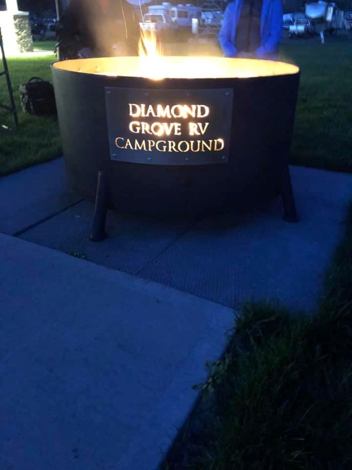 Cylindrical fire pit with Diamond Grove RV Campground sign in outdoor campground setting.