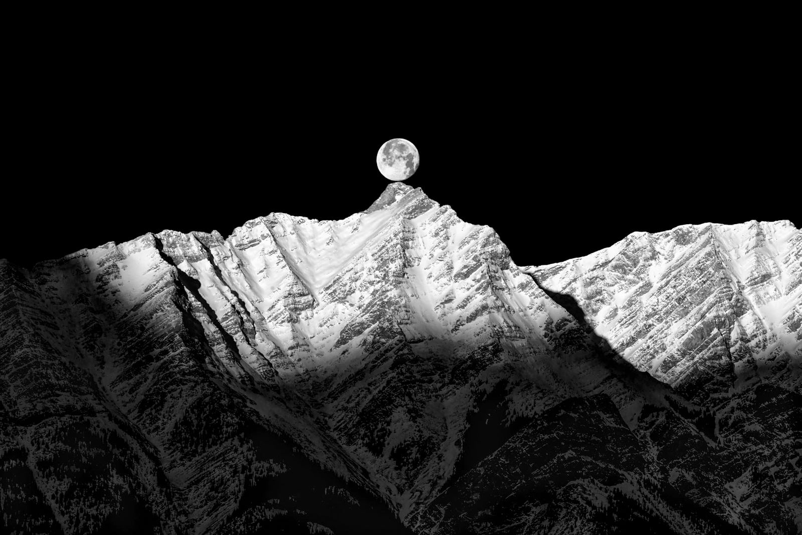 A black and white photo of the moon on a black sky over the peaks of a mountain range.