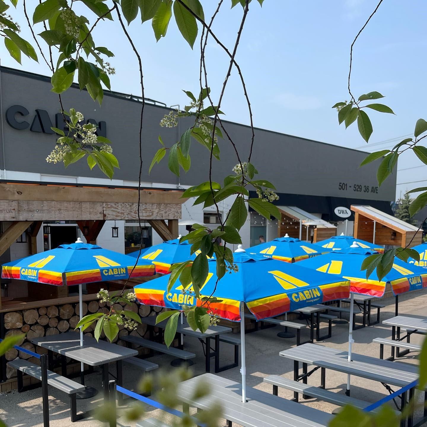 Patio umbrellas with Cabin Brewing branding, benches, and industrial building facade in Calgary.