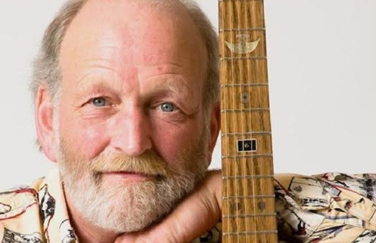 Bearded person in patterned shirt resting chin on hand beside guitar neck.