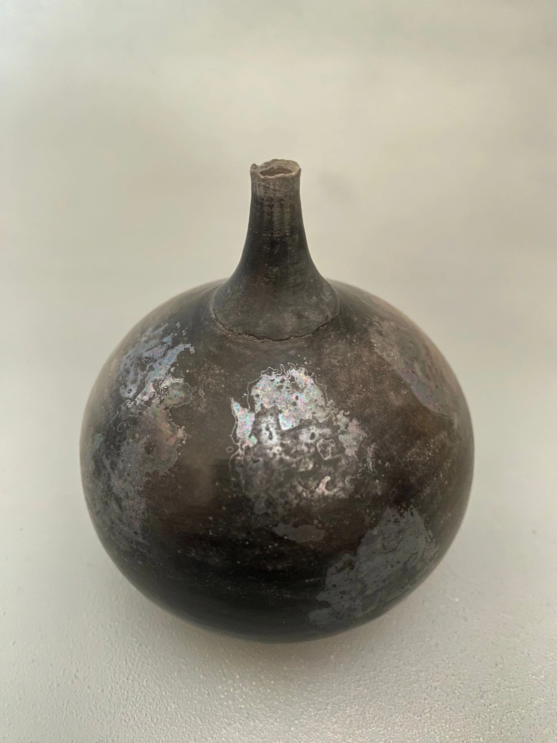 Handmade round ceramic vessel with a narrow neck and dark, textured glaze on a light background.