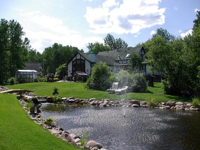 Lush garden with pond and fountain in front of a large house on a sunny day.