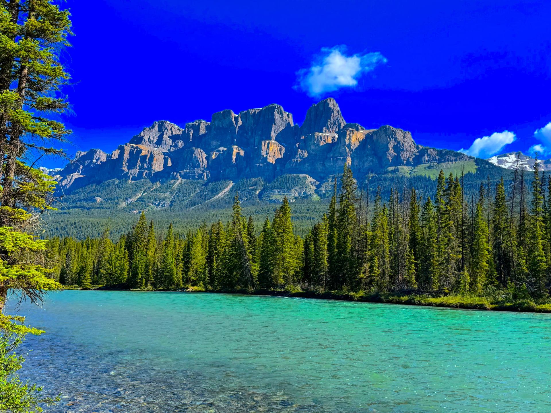 Turquoise river and evergreen forest beneath rugged mountains and a vivid blue sky.