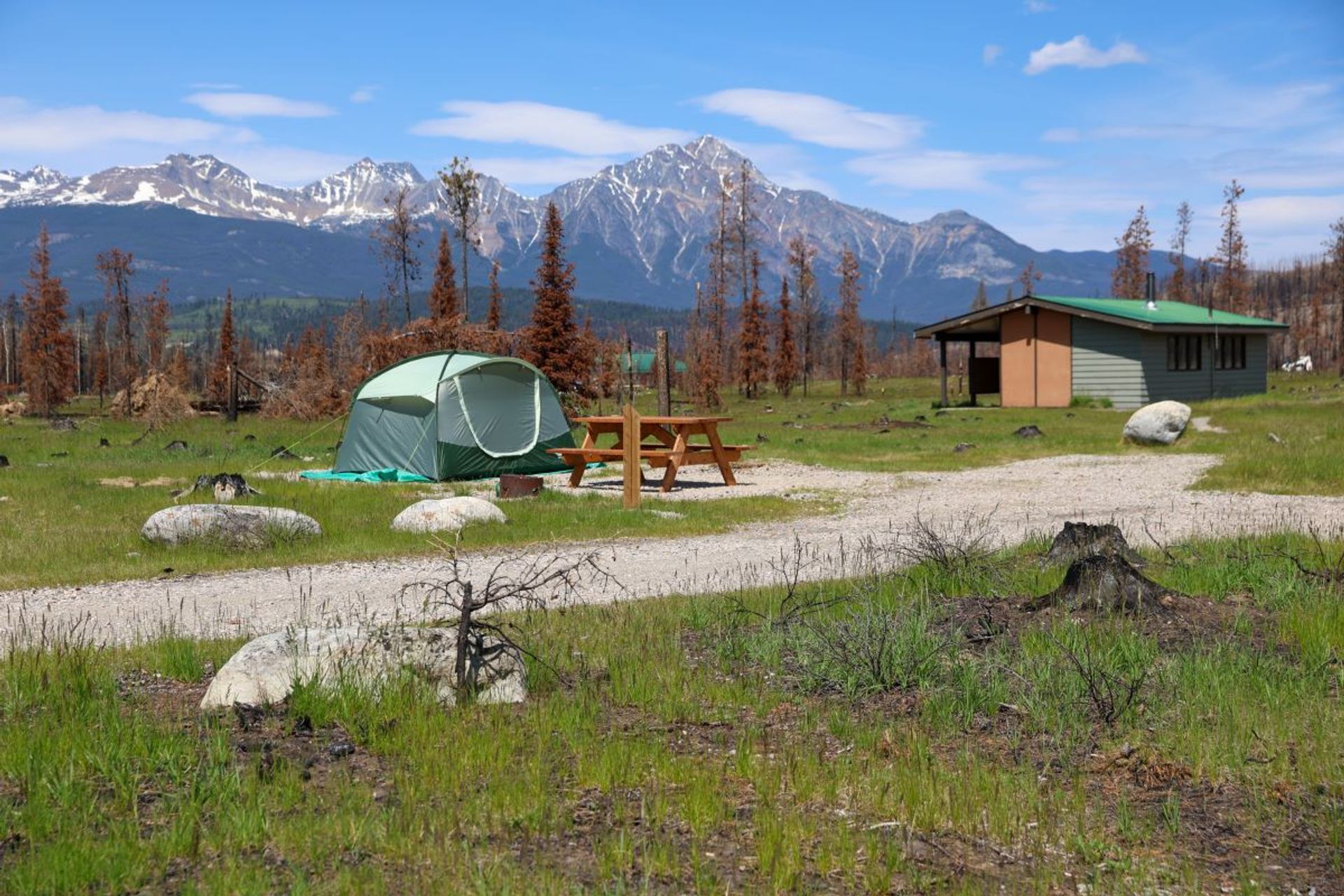 Green tent and picnic table on grassy campsite with mountains and cabin in background.