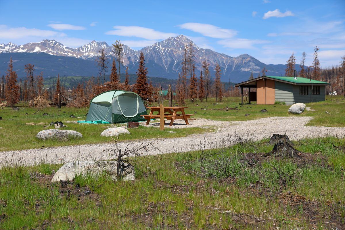 Green tent and picnic table on grassy campsite with mountains and cabin in background.