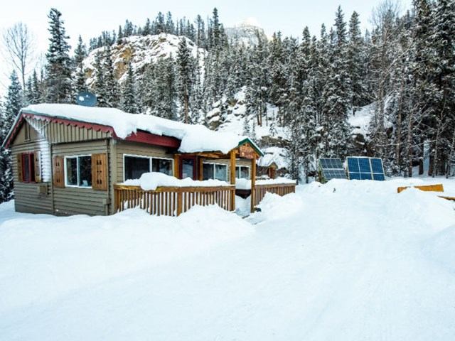 Snow-covered cabin with red trim, solar panels, and forested mountain backdrop.