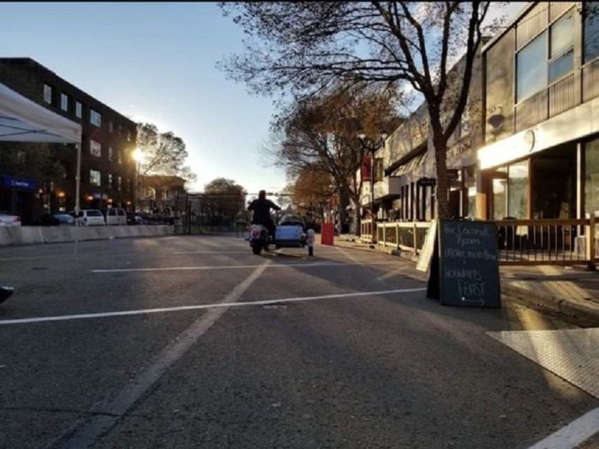 People walk and bike along Ross Street with shops and trees.