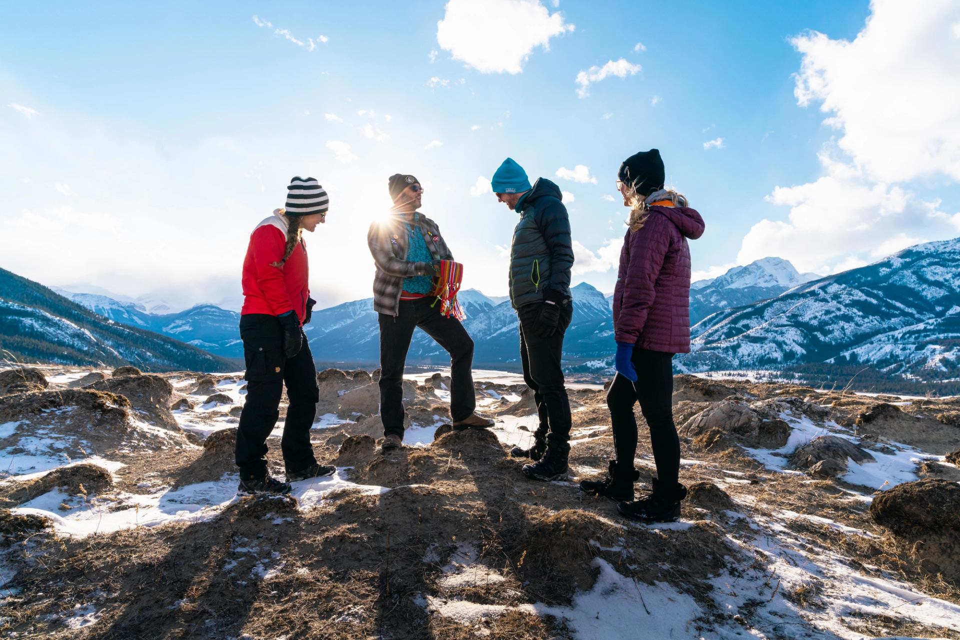 Four in winter gear stand on snowy mountain, sunlight shining through as they enjoy the view.
