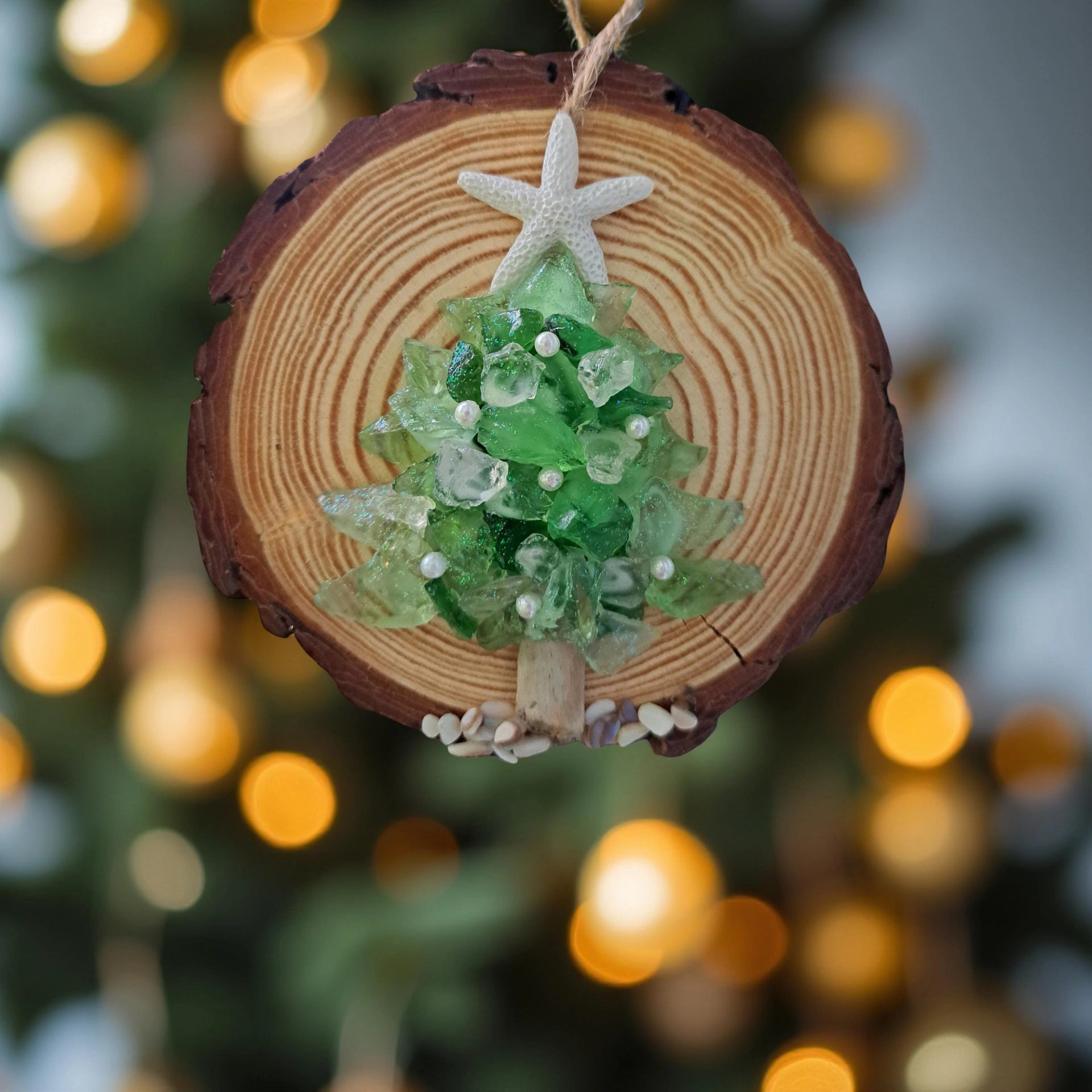 Wood slice ornament with glass Christmas tree and starfish, lights blurred in background.