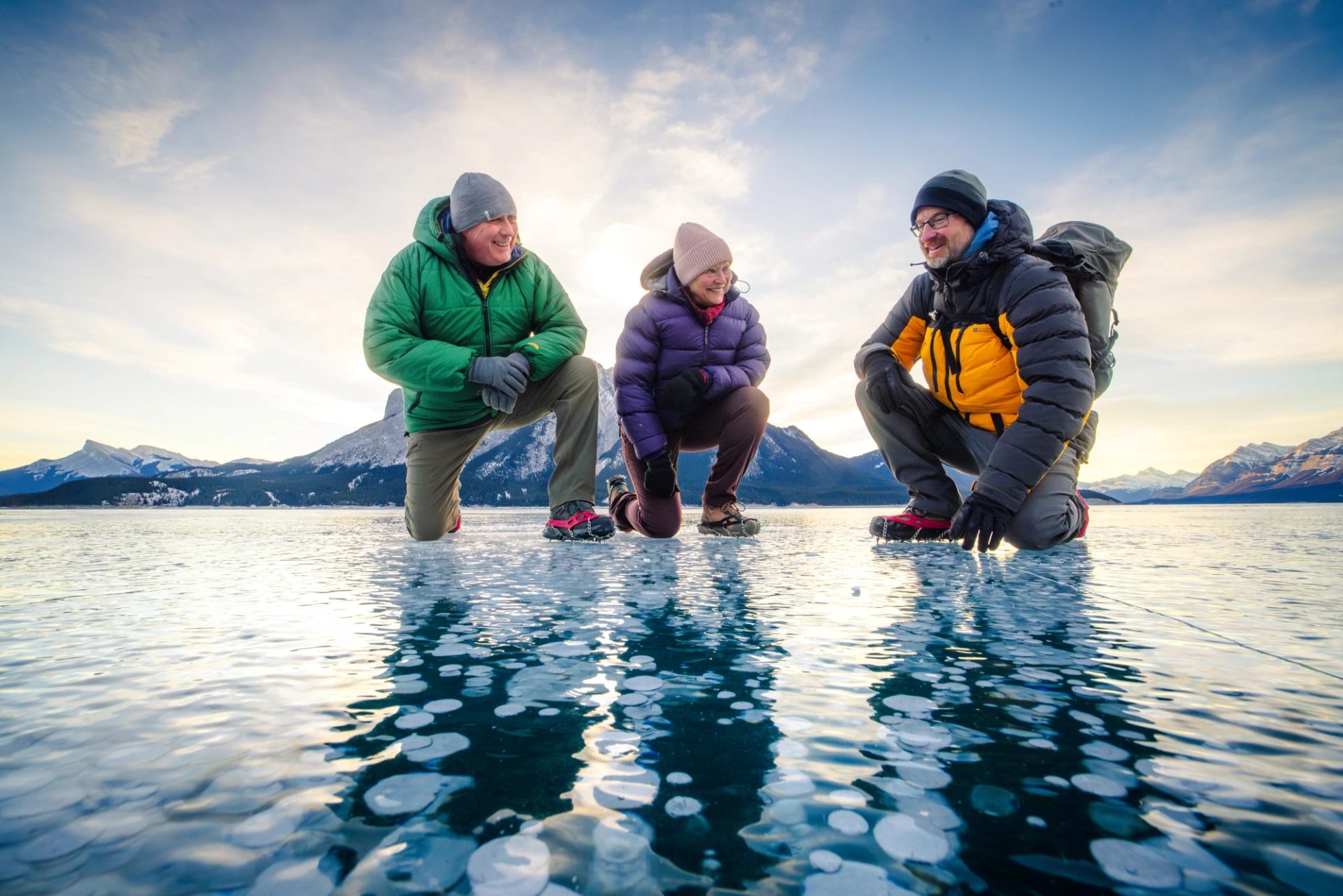 Three people kneel on a crystal‑clear frozen lake, surrounded by snowy peaks and soft winter sunlight.