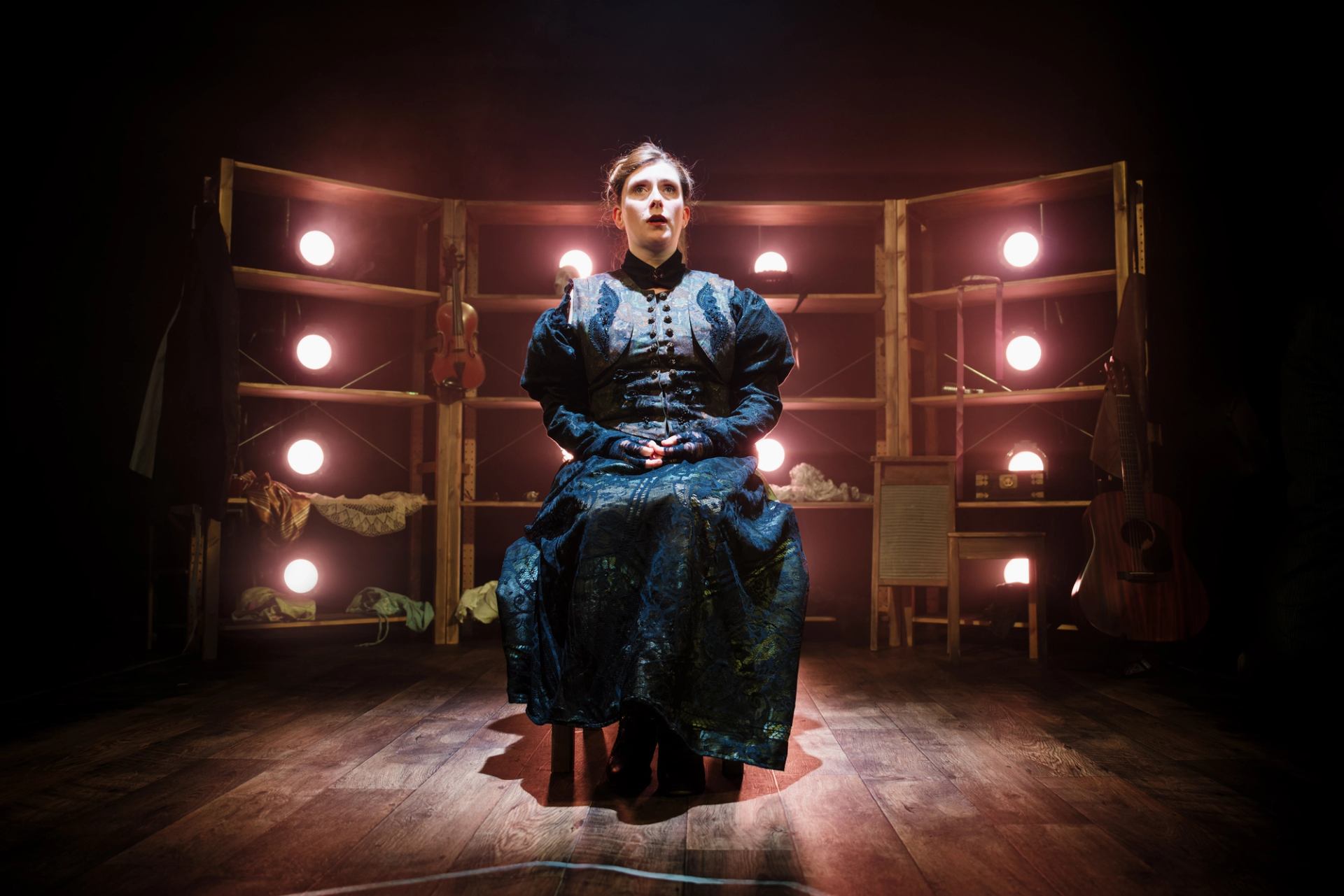 Stage performer in dark period costume sits under warm lights, instruments on shelves behind.
