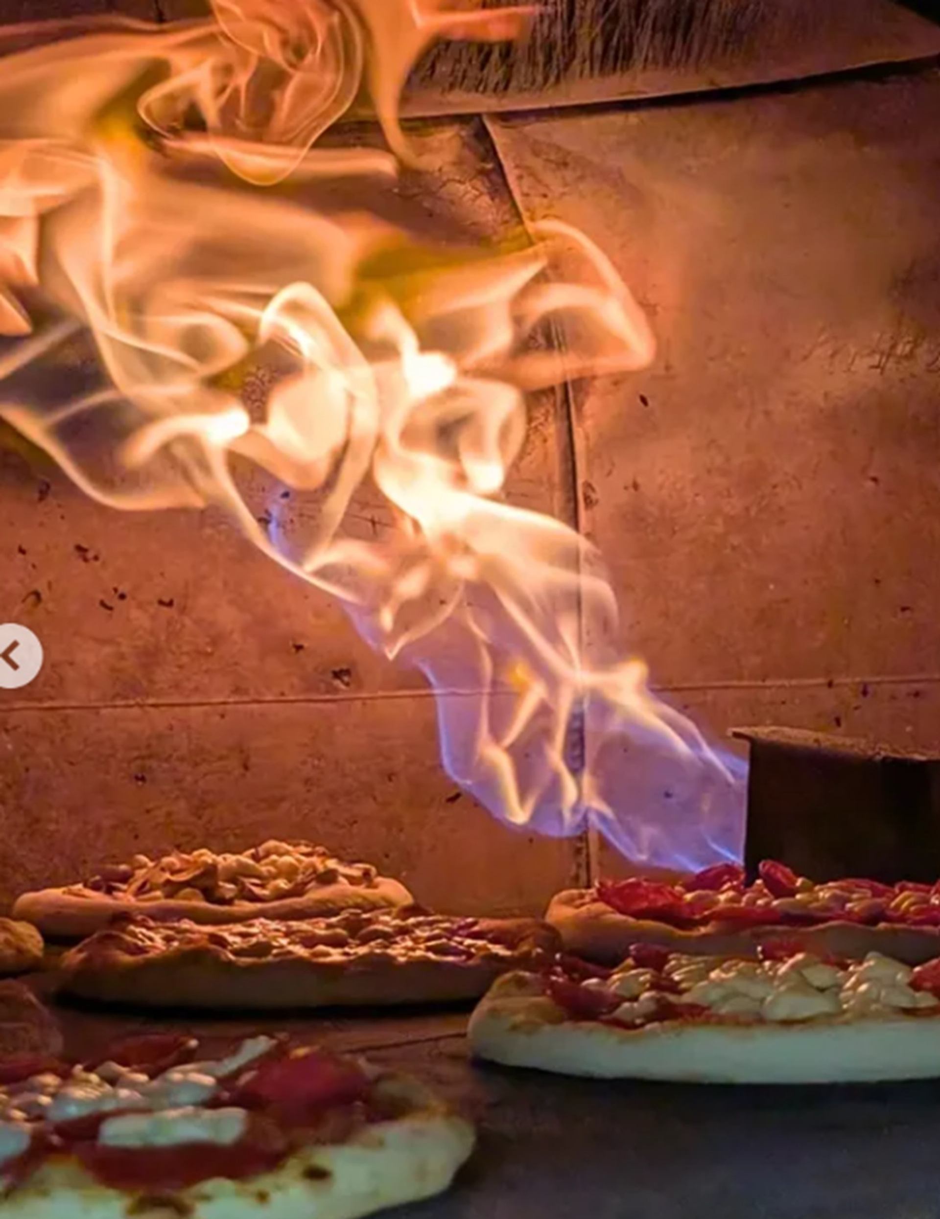 Orange and blue flames rise above pizzas inside a brick oven.