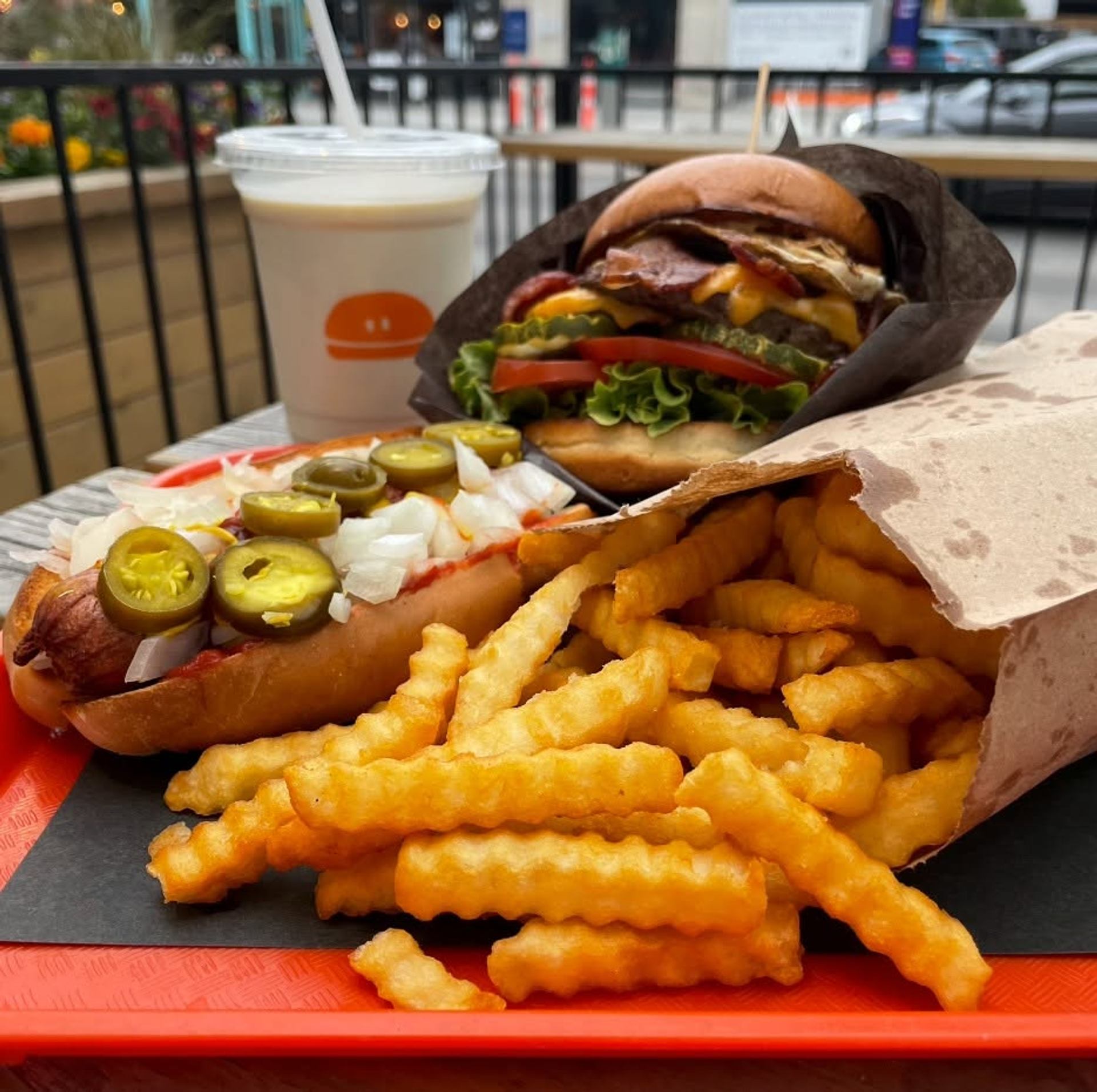 A burger, hot dog, crinkle-cut fries, and milkshake are served on a bright tray outdoors.