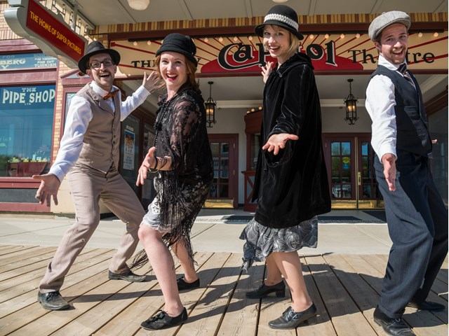 Dancers pose outside Capitol Theatre entrance, wearing costumes and hats on wooden sidewalk.