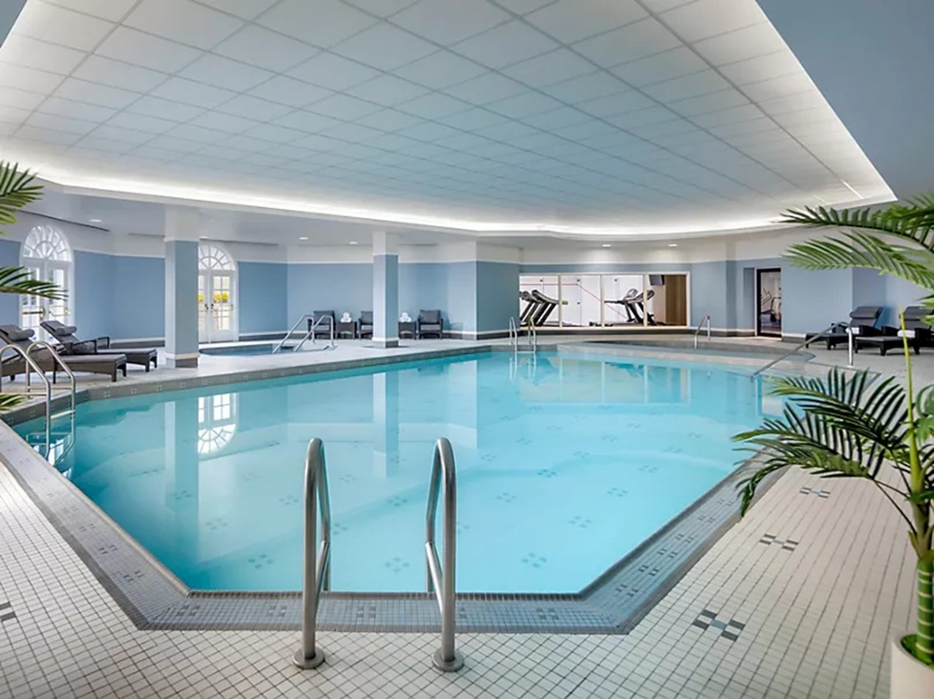 Indoor swimming pool with tiled deck, lounge chairs and adjacent fitness room with treadmills.
