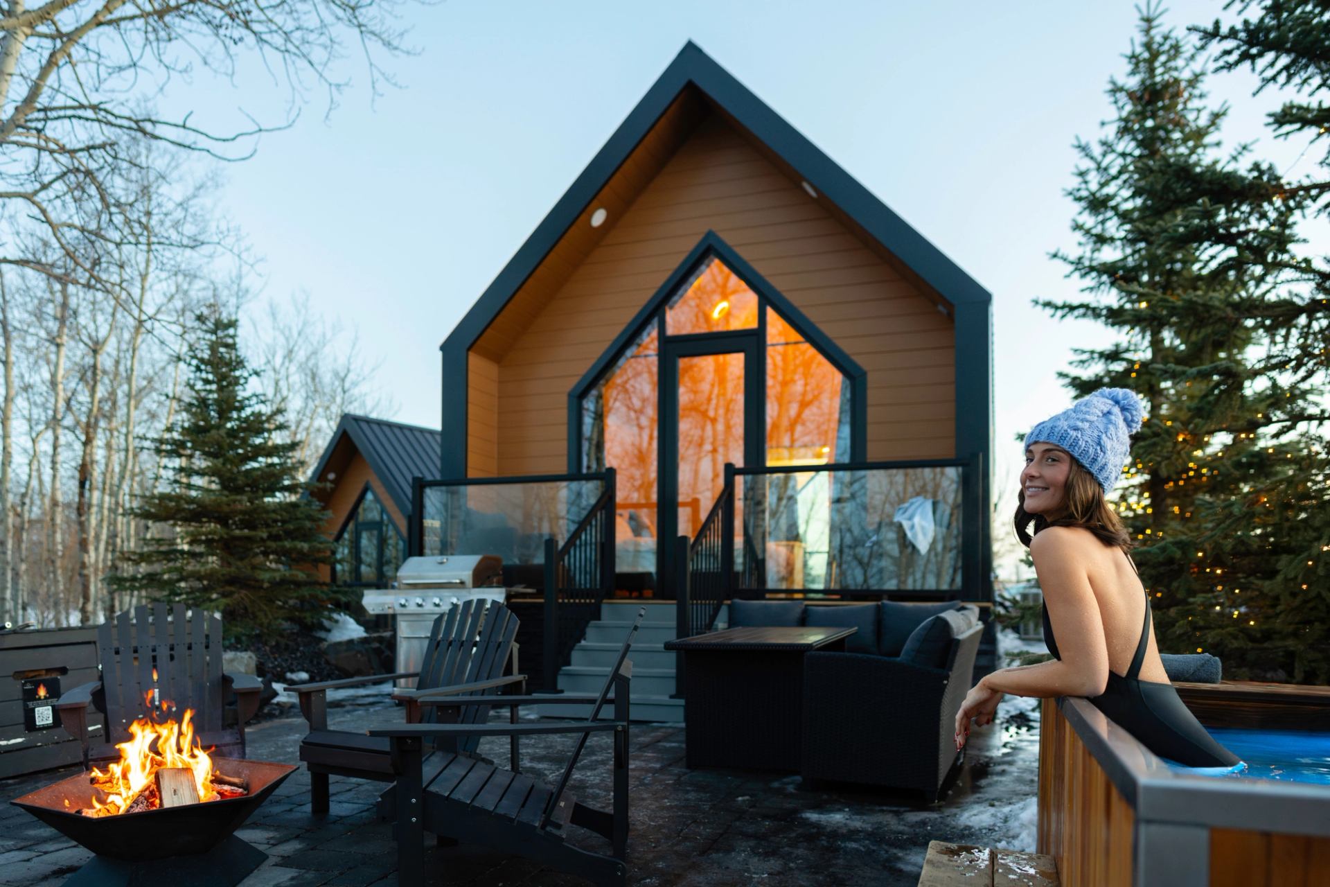 Outdoor cabin patio with firepit, hot tub and person in swimsuit wearing knit toque.