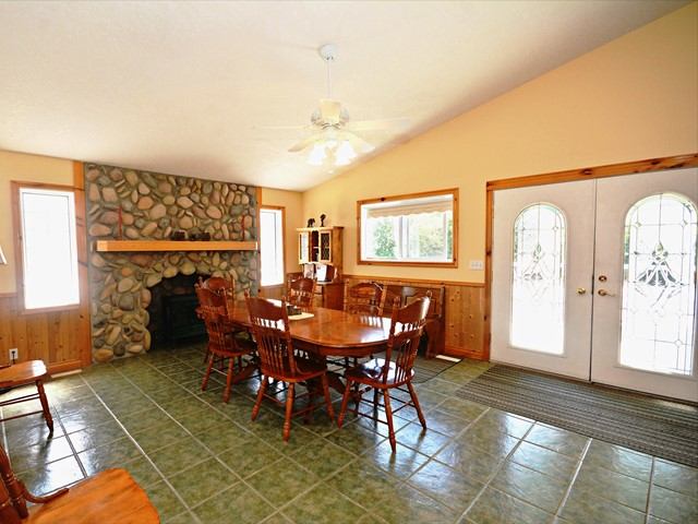 Cozy dining room with wooden table, stone fireplace, large windows, and a bright entrance door nearby.