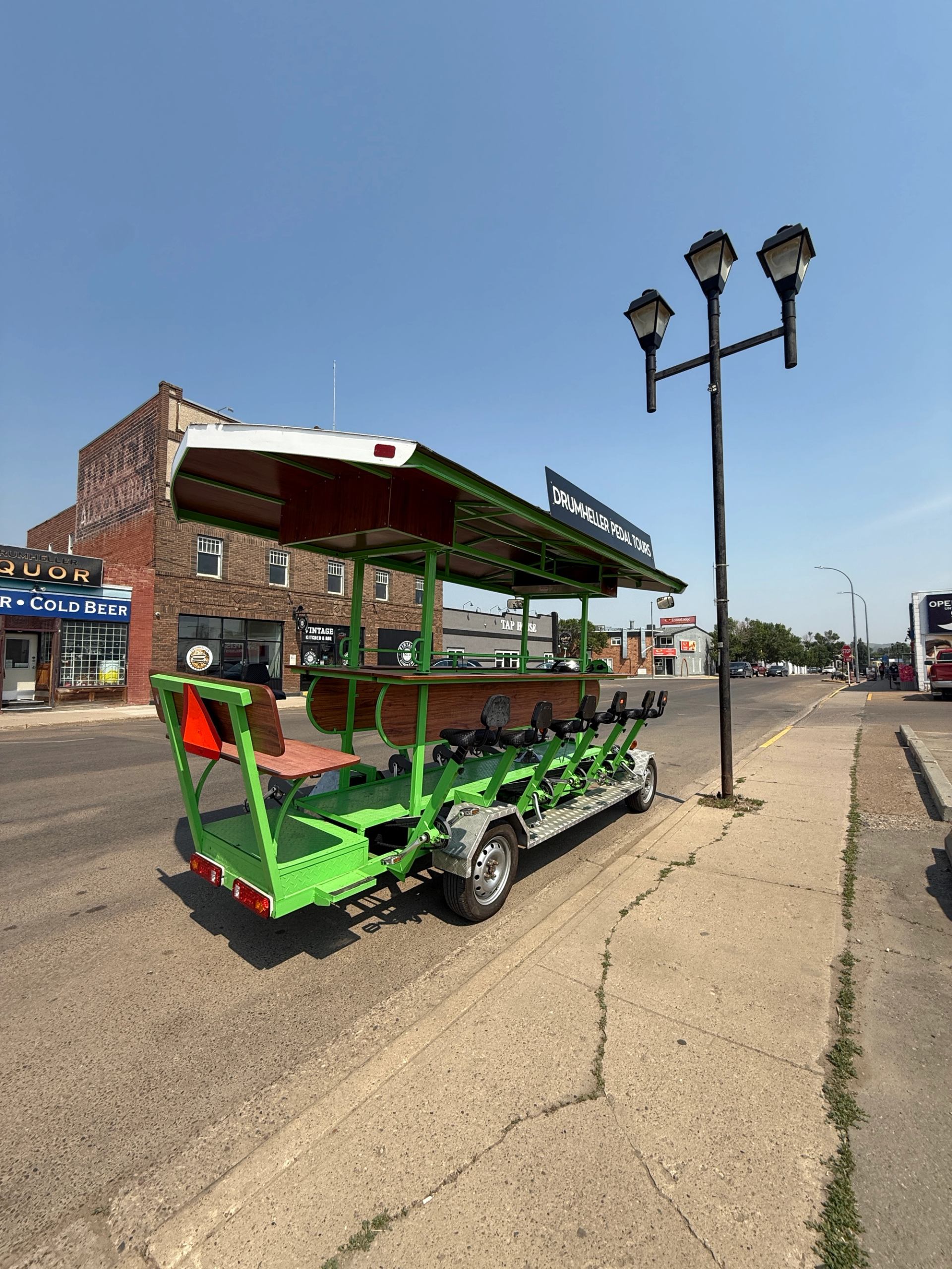 Green pedal tour trailer with open roof, street lamps, and storefronts in Drumheller.