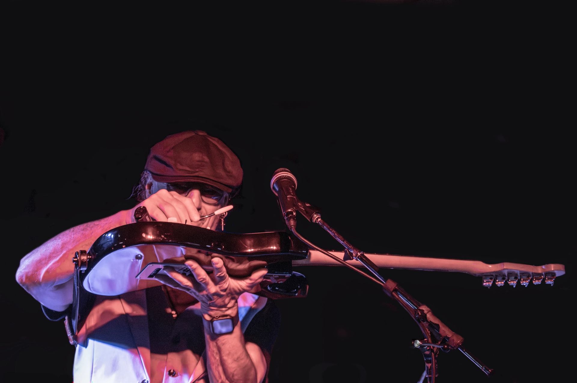 Musician in a cap holds an electric guitar horizontally beside a microphone.