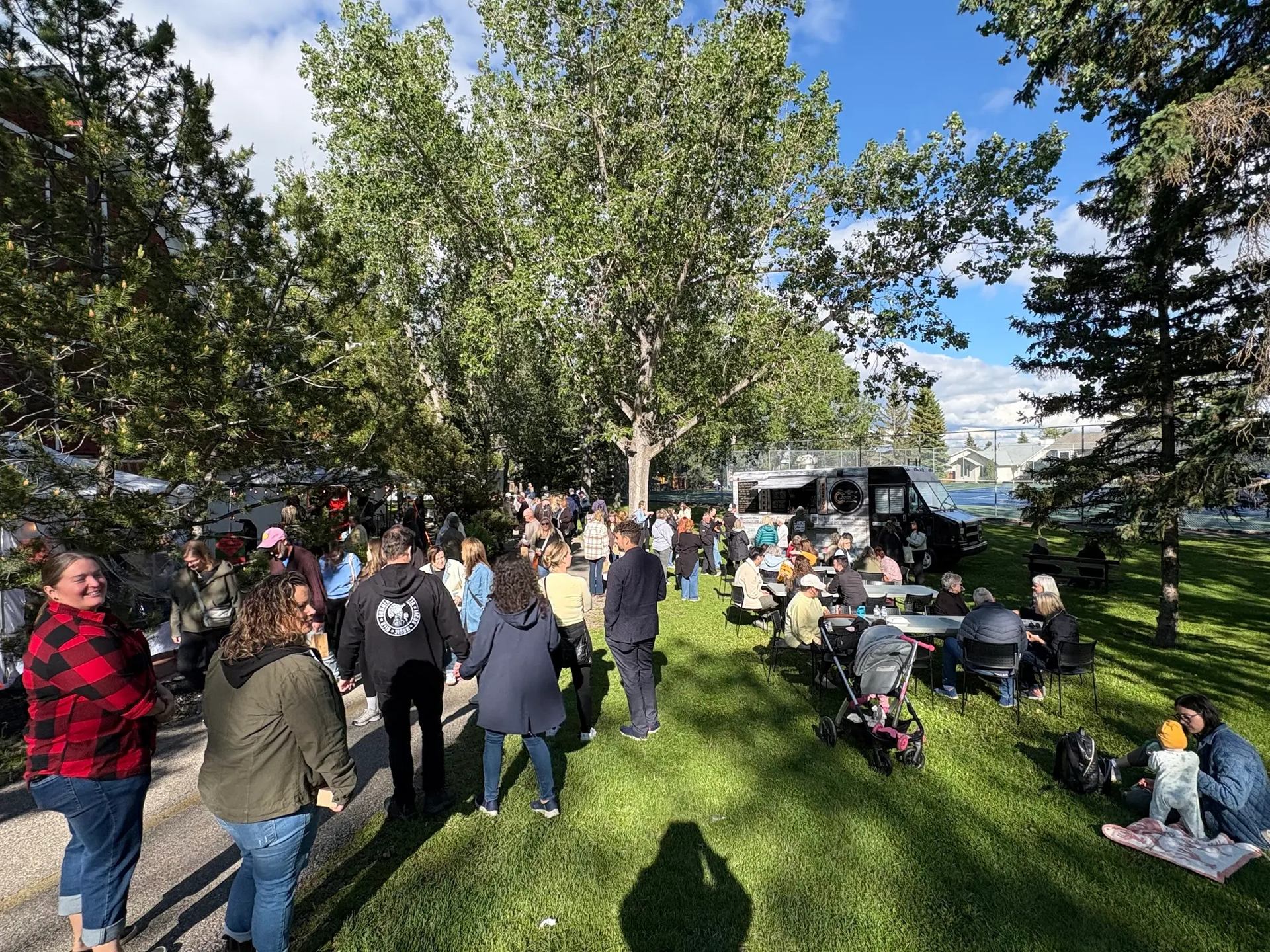 Crowd gathers in a tree-filled park with food truck, tables and strollers.