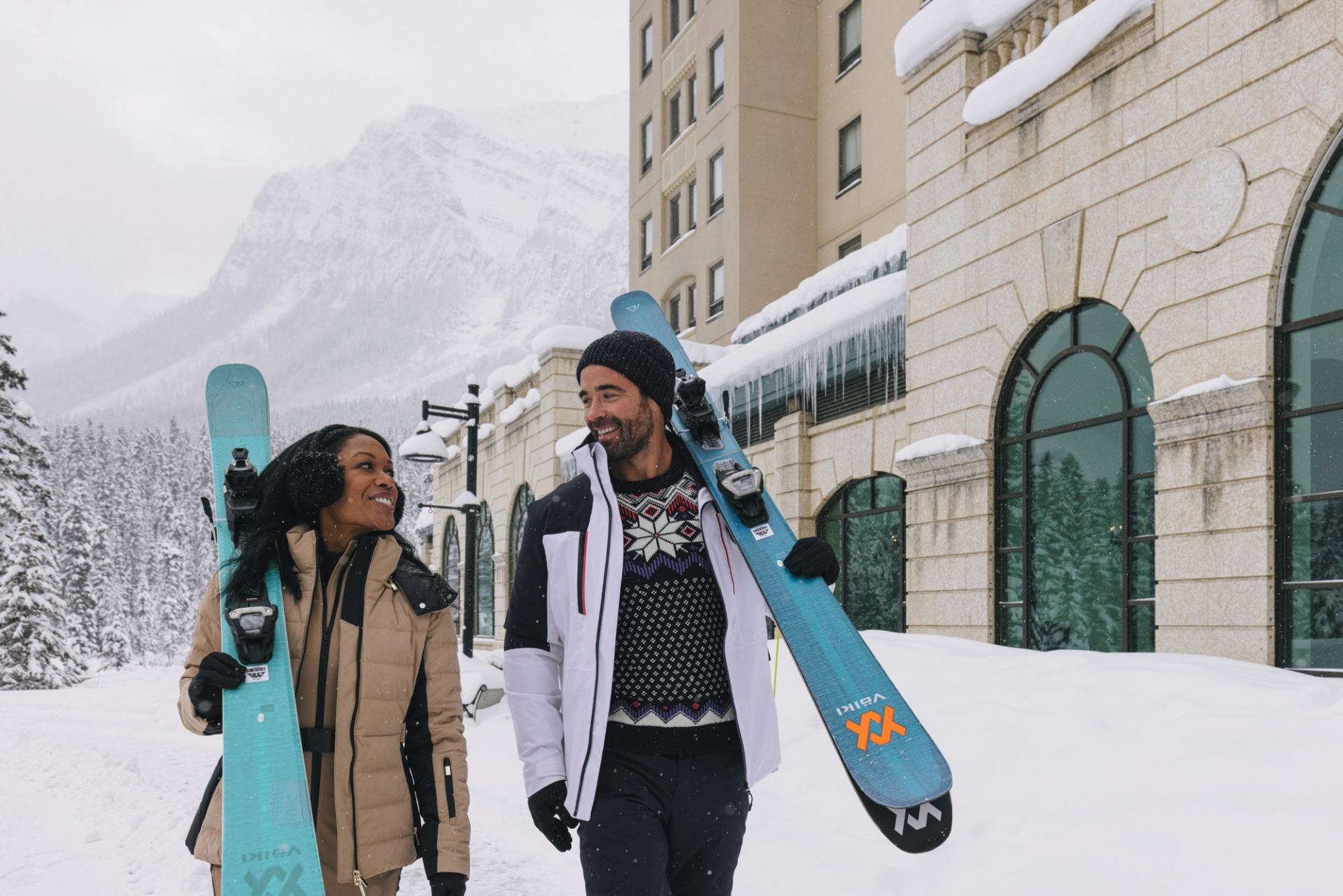 Skiers carrying skis walk past snowy hotel building with mountains and trees behind.