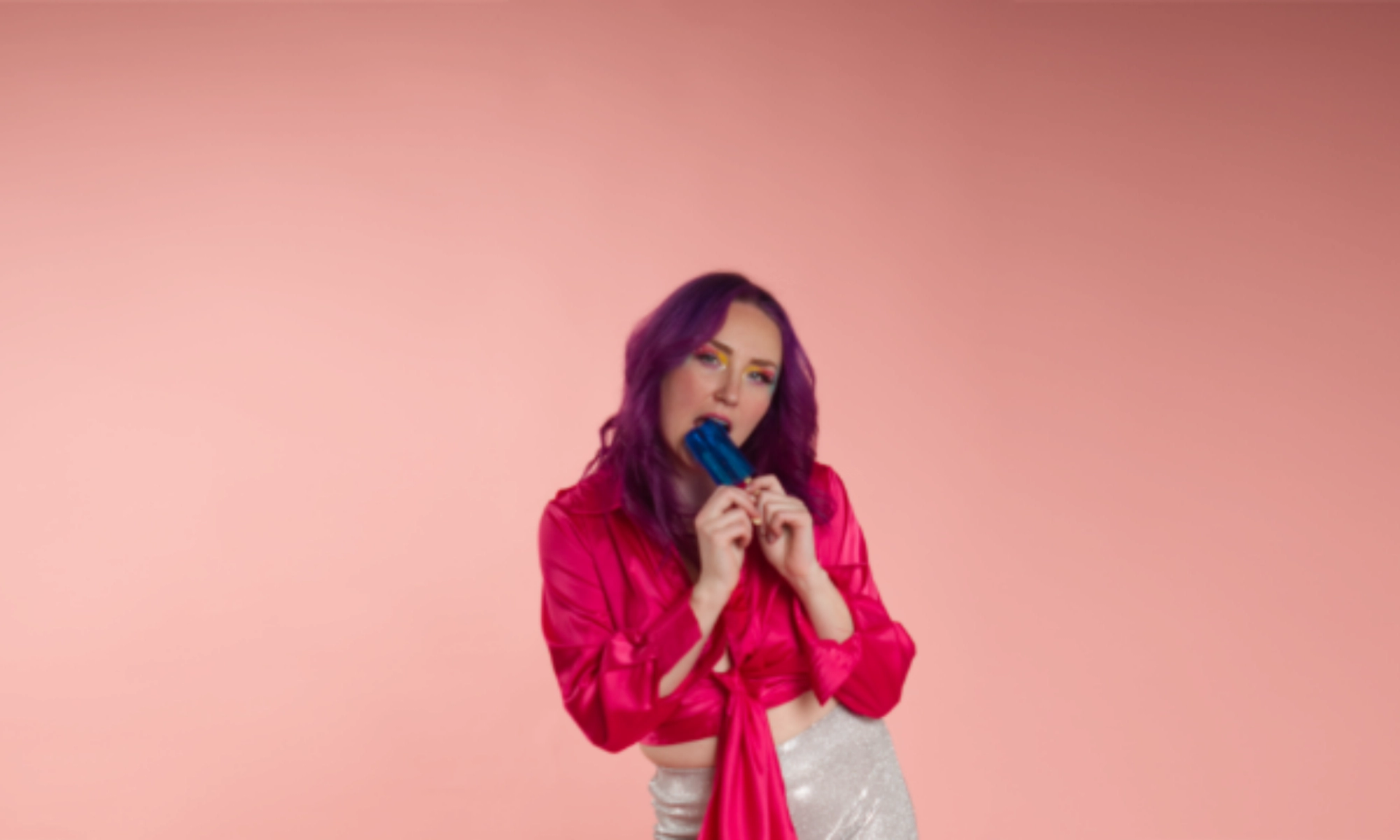 Person with purple hair holds blue popsicle against mouth before pink background.