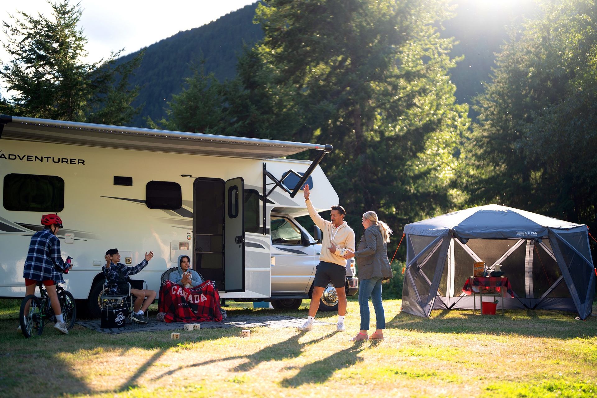 Group camping near RV and tent in a scenic forested area with mountains in the background.