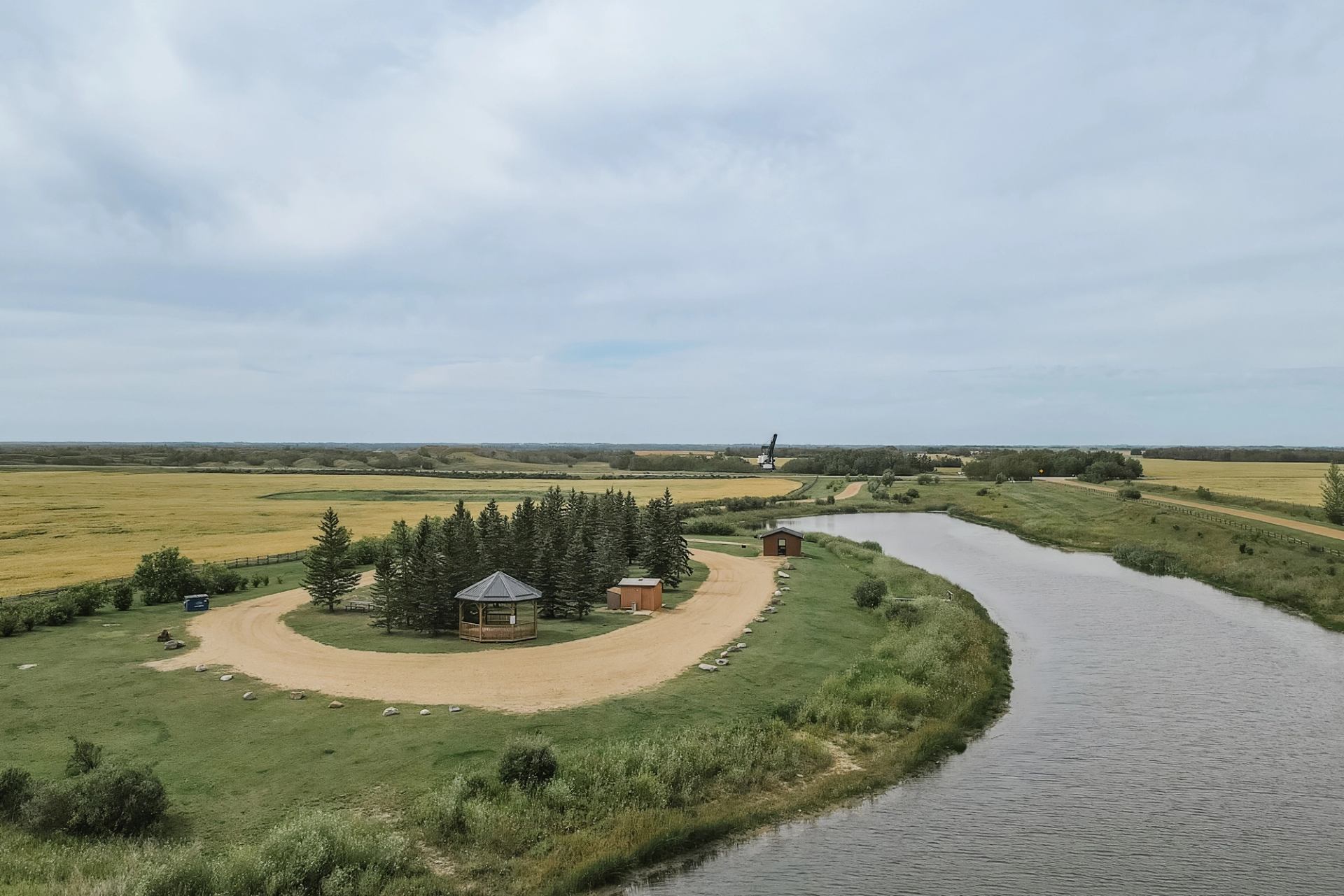 Riverside park with gazebo, trees and gravel road beside wide, curving waterway.