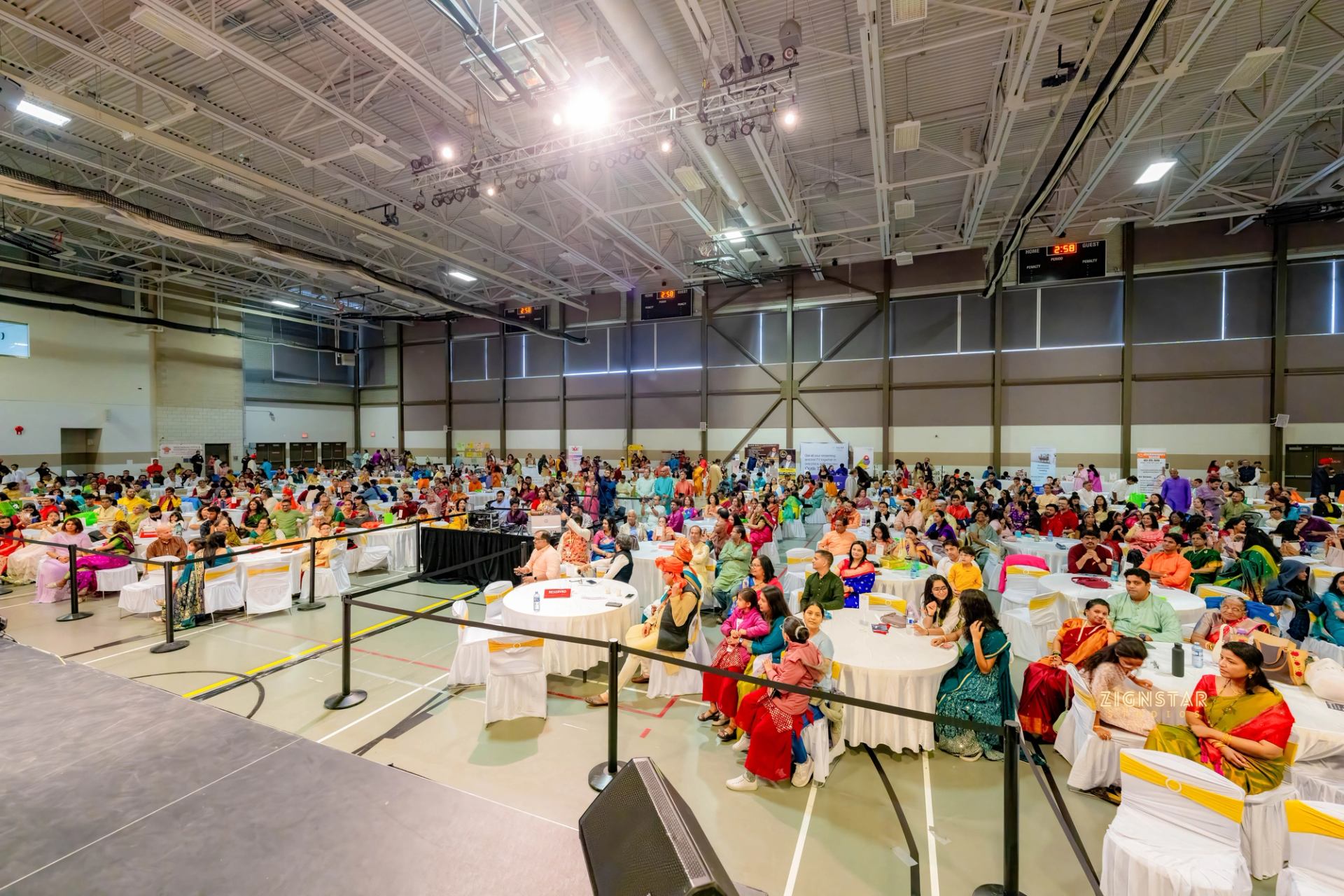 Large crowd in colourful clothing sits at round tables inside a gymnasium.