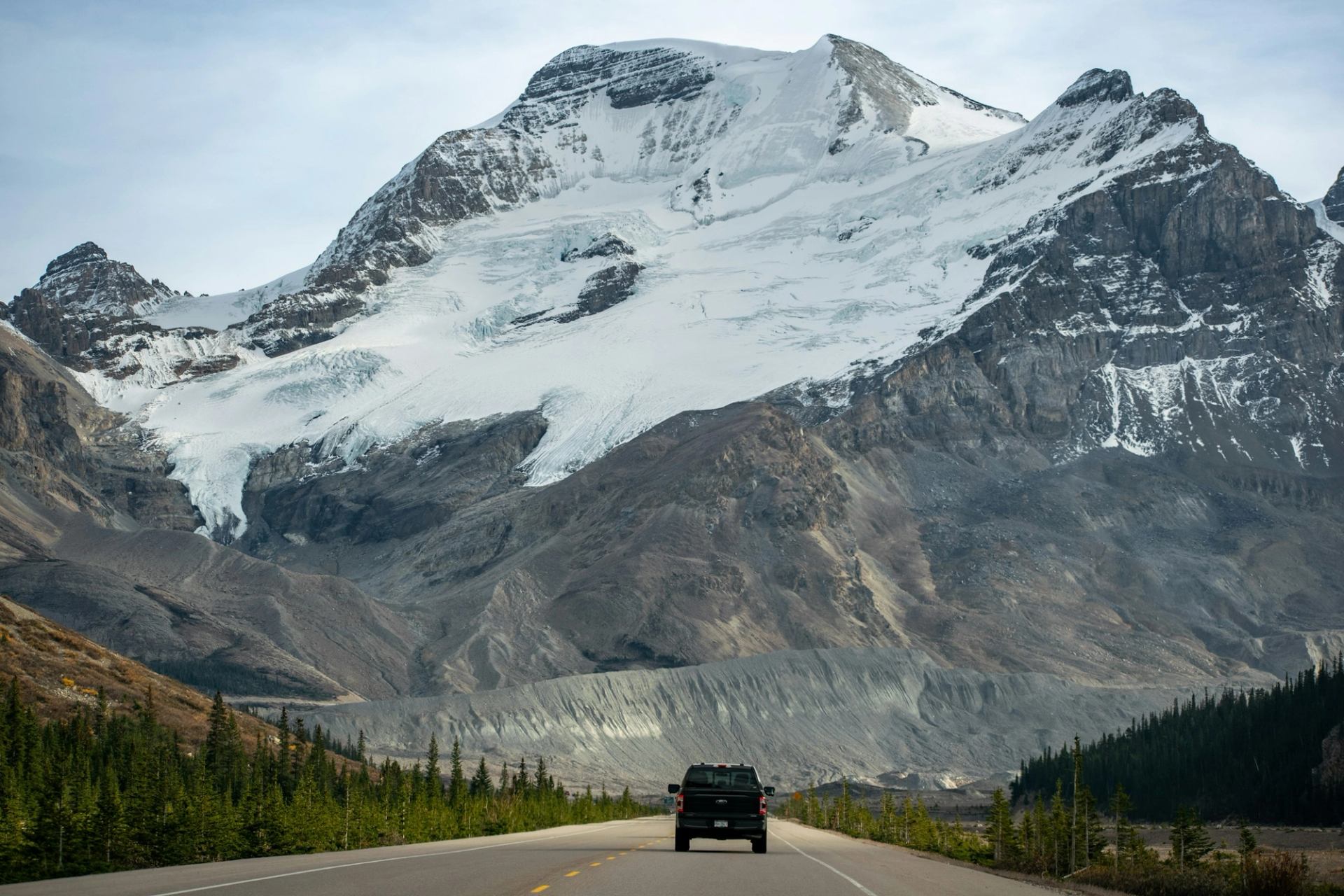 Pickup truck drives toward a snow-covered mountain glacier above forested valley.