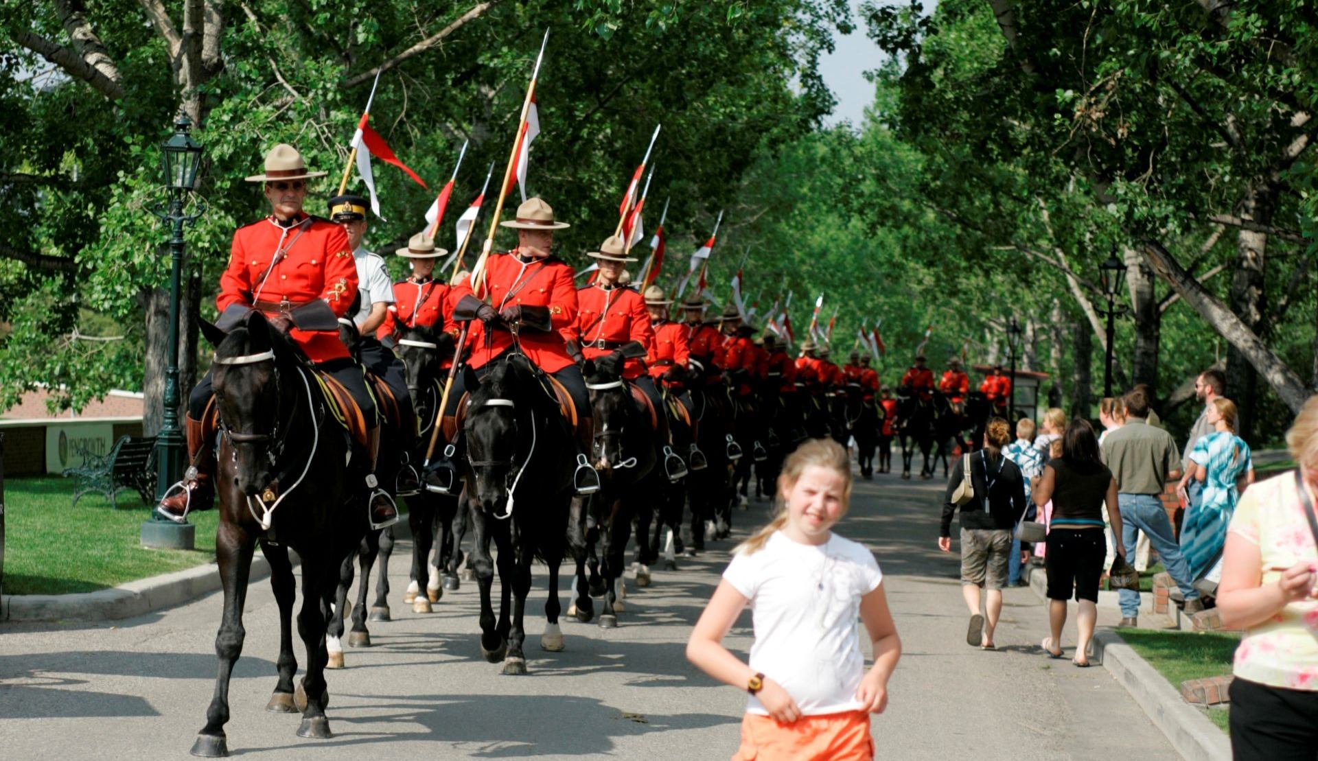 RCMP officers in red uniforms ride horses in formation during a parade as people watch nearby.