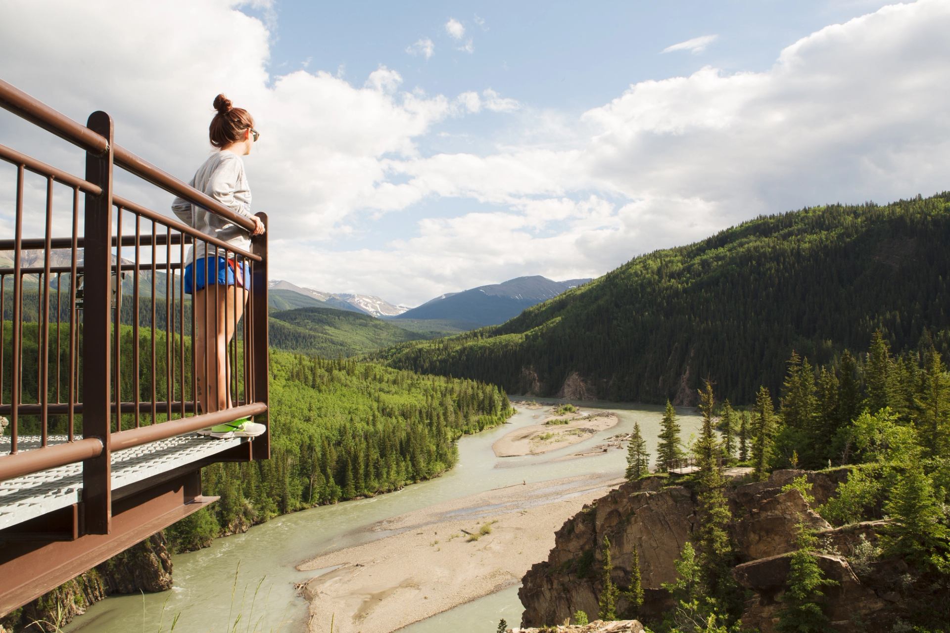 Person on a lookout platform views a winding river through forested mountains.