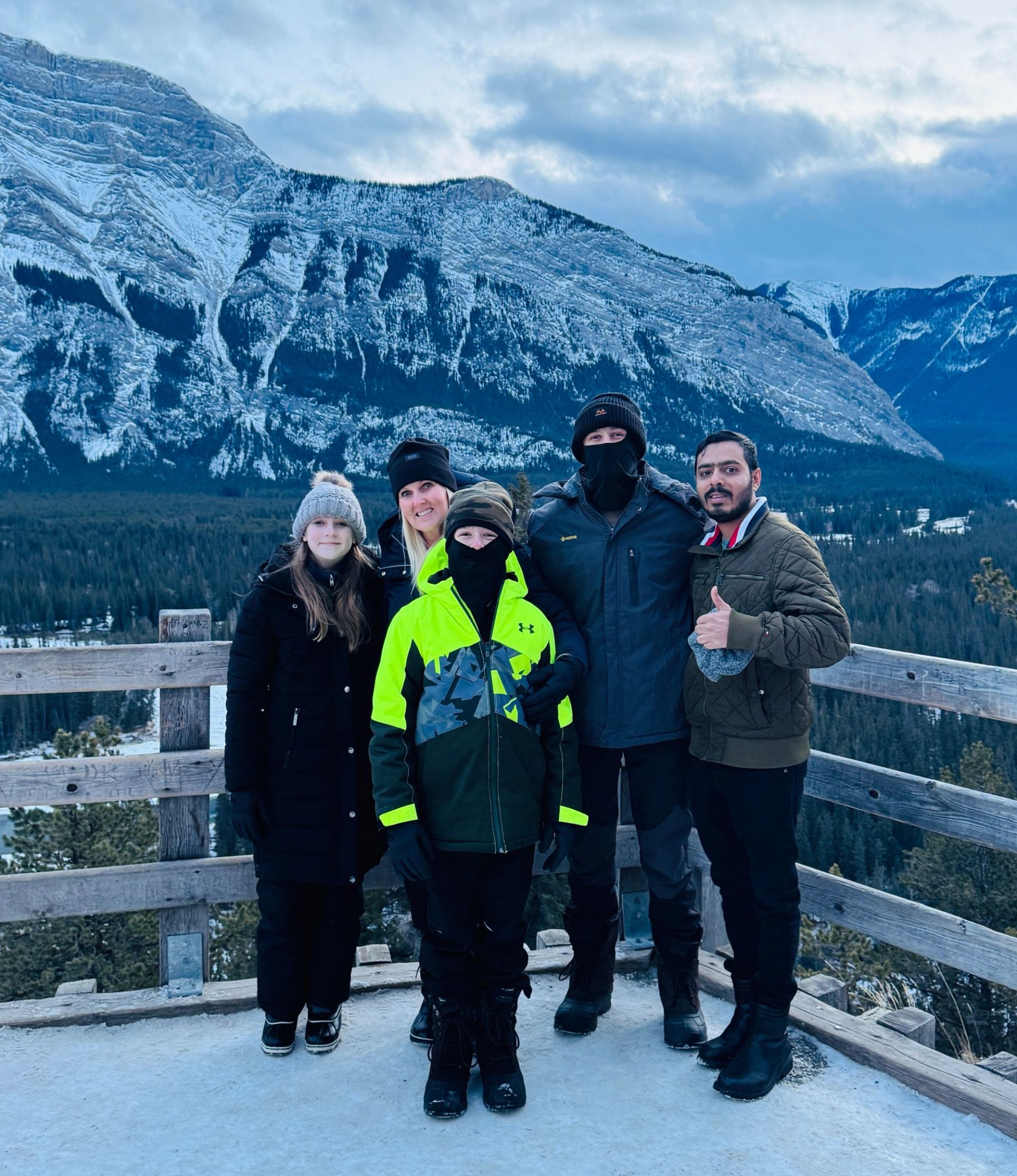 Group of people in winter clothing on wooden lookout deck with snowy mountains behind.