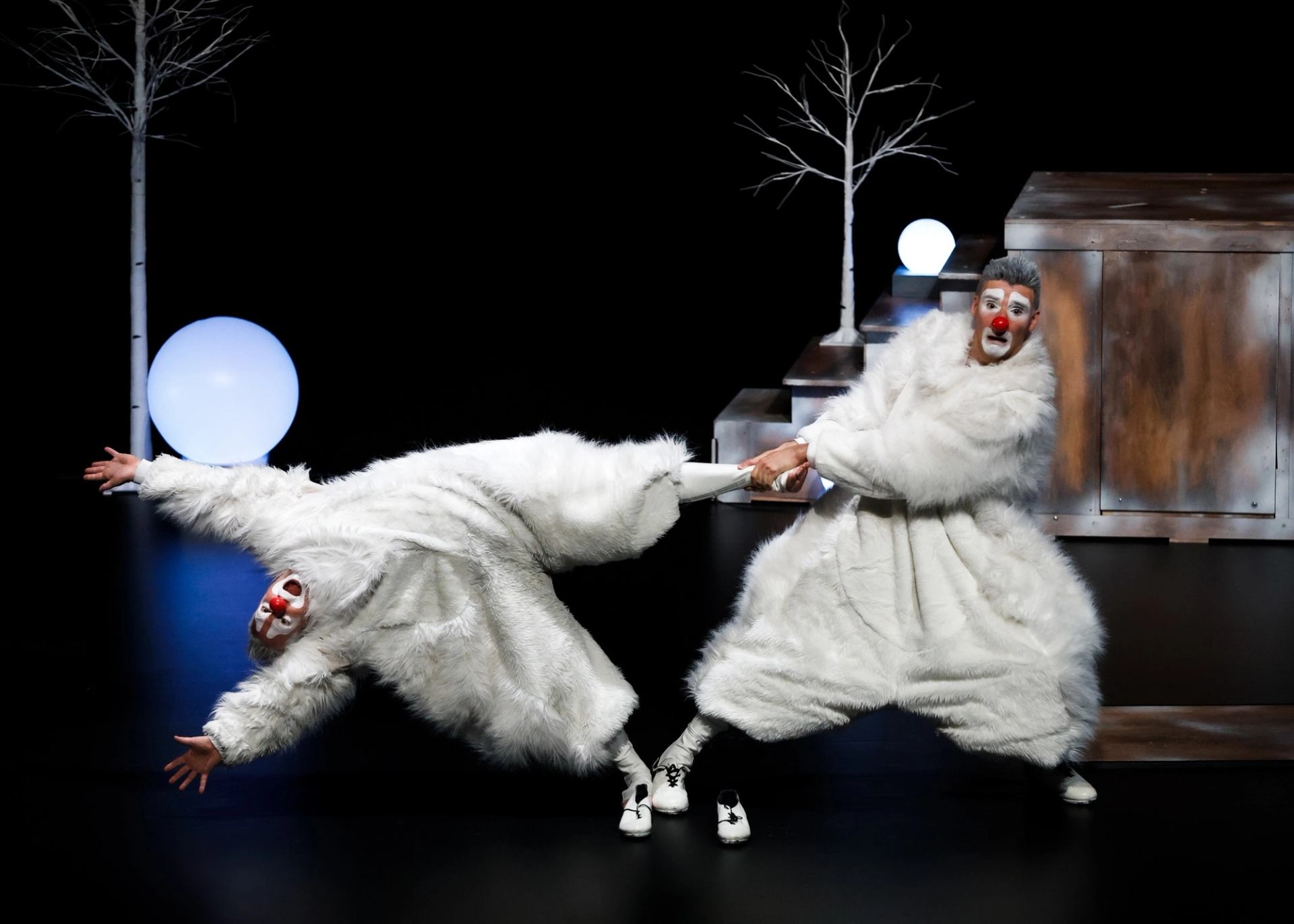 Two clowns in white furry costumes tug arms onstage beside bare trees and glowing orbs.