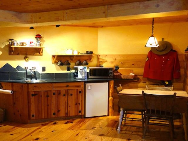 Rustic wood kitchen with shelves, mini‑fridge, table, and a red jacket hanging on the wall.