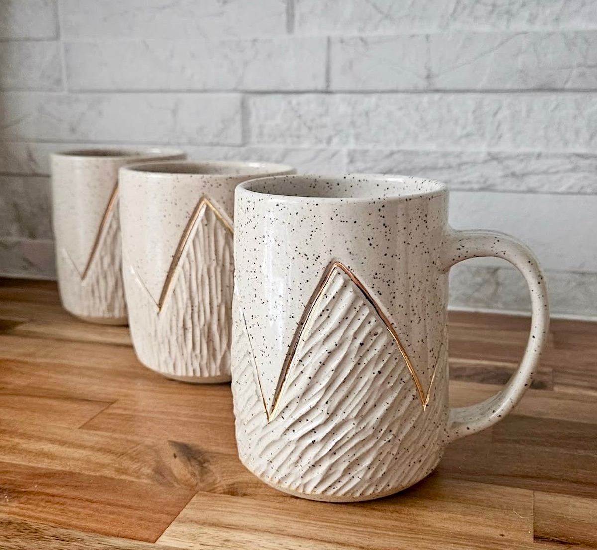 Speckled ceramic mugs with carved mountain pattern on wooden counter before tiled wall.