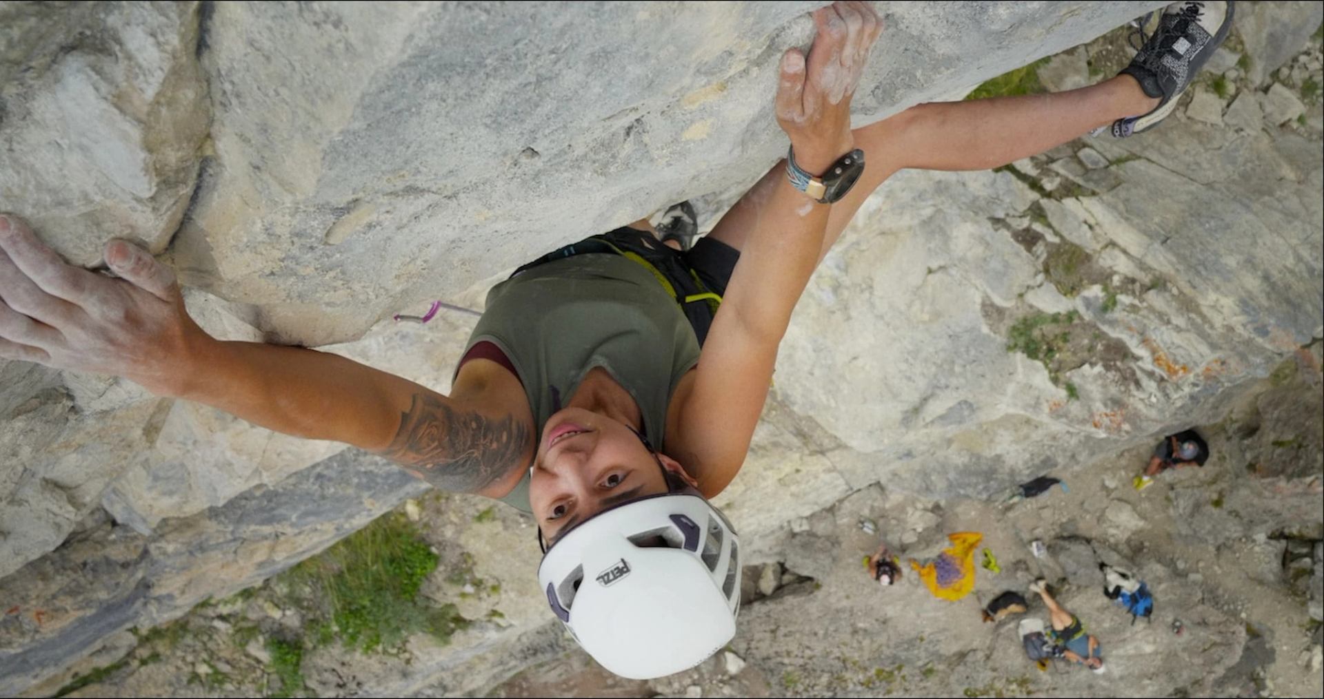 Rock climber wearing helmet and harness ascends steep cliff, with people visible below.
