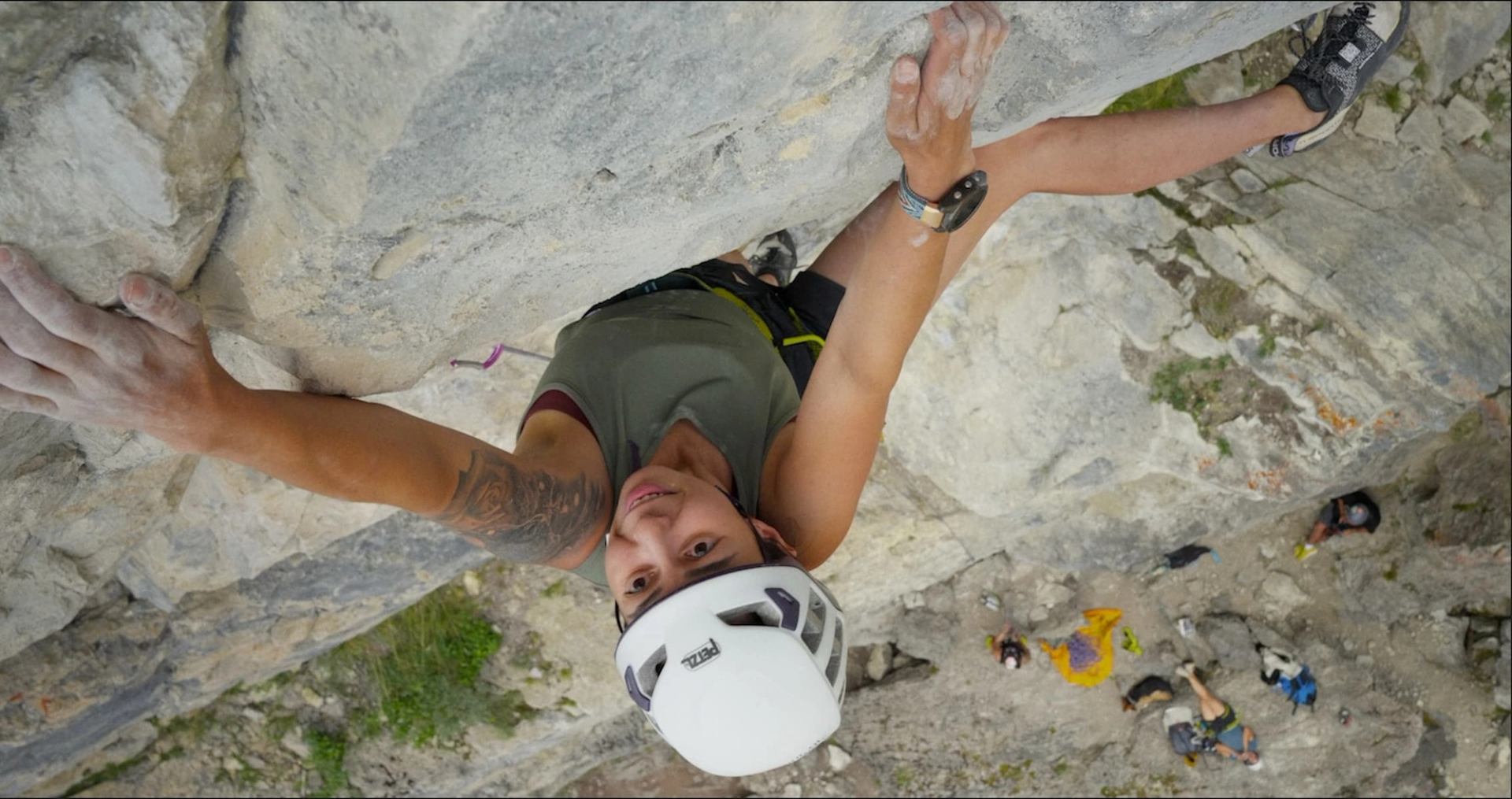 Rock climber wearing helmet and harness ascends steep cliff, with people visible below.
