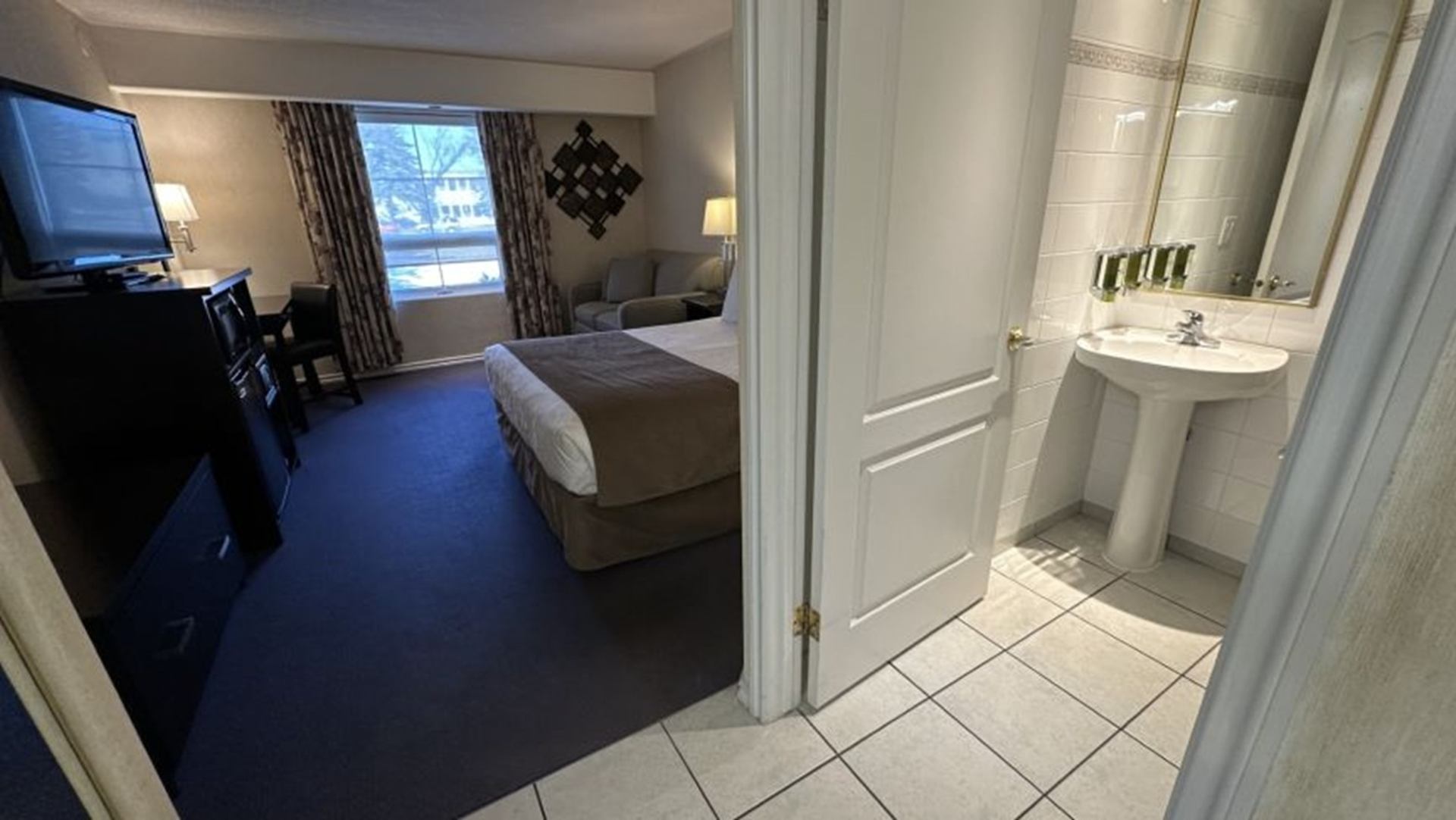 Hotel room with bed, sofa and desk beside open door to tiled bathroom.