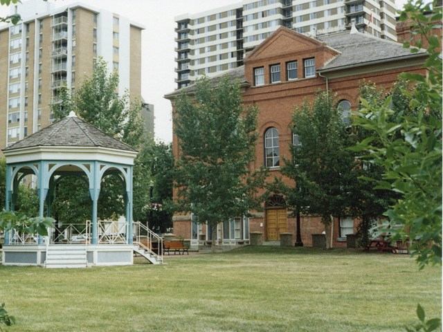 Red brick building with arched windows near gazebo and high-rises in a green urban park.
