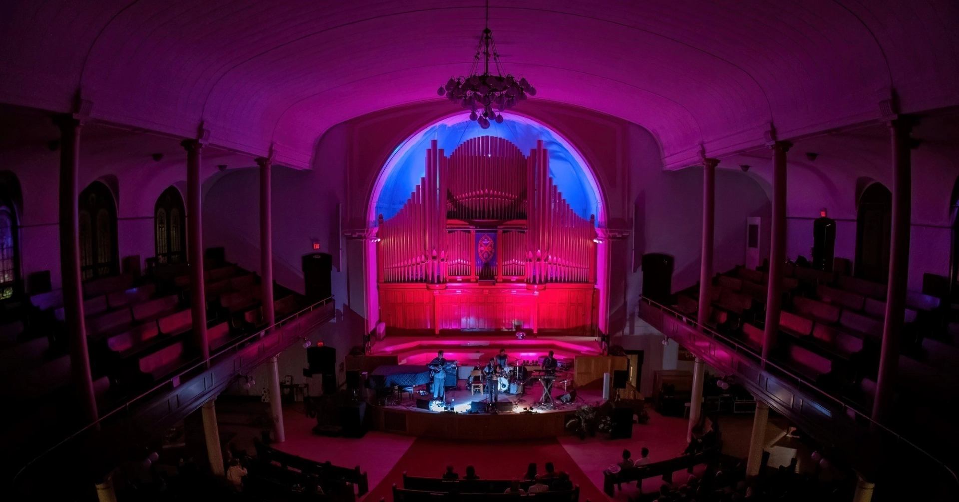 Concert hall stage with organ pipes and musicians under purple and blue lighting.