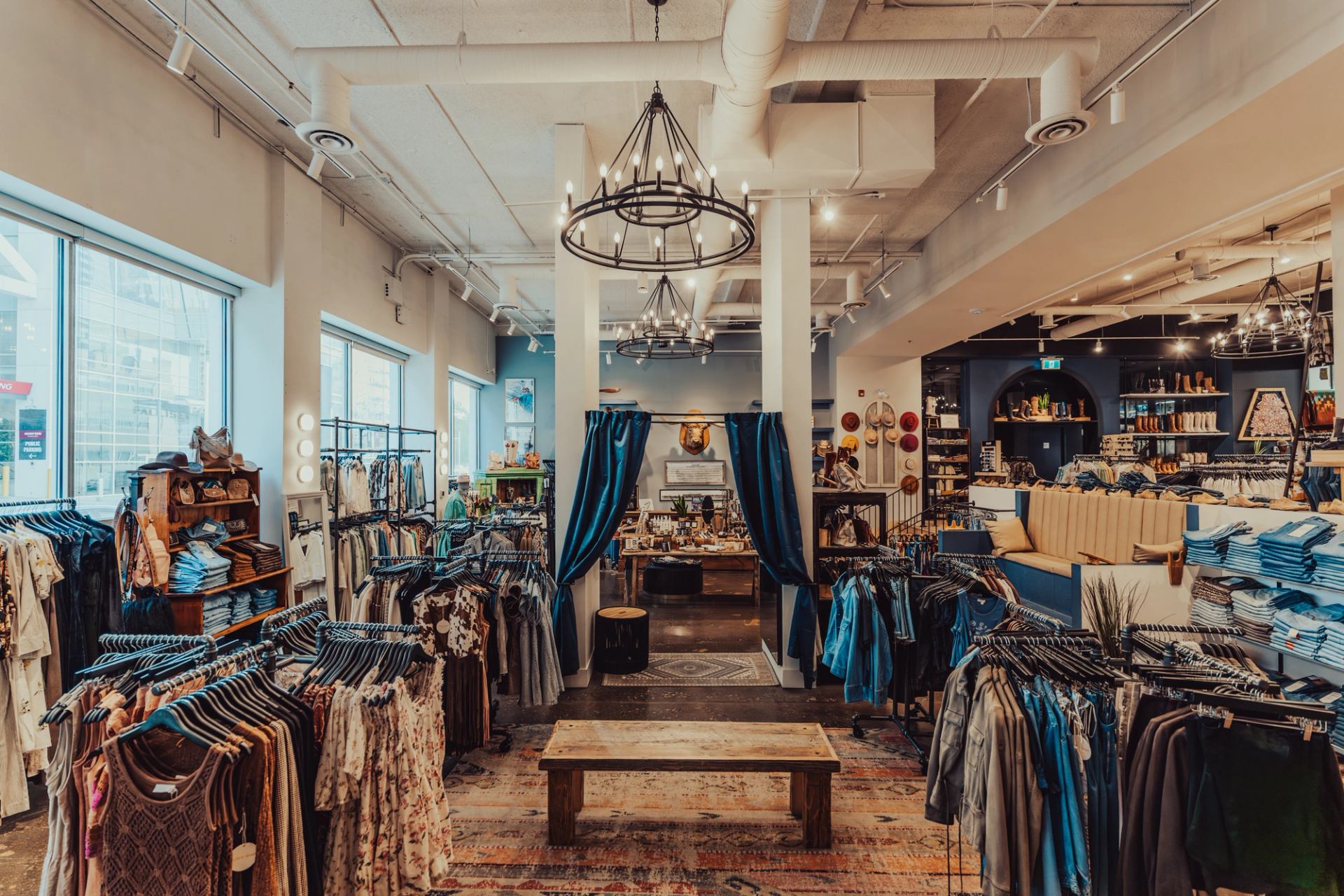 Clothing boutique interior with racks of garments, folded denim, chandeliers and seating area.