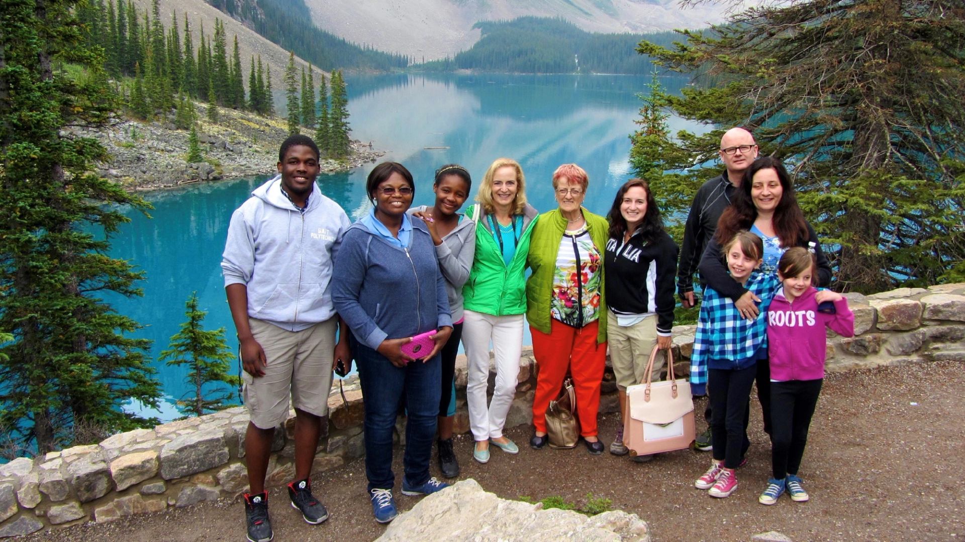Group of people smiling in front of a turquoise lake surrounded by pine trees and mountains.