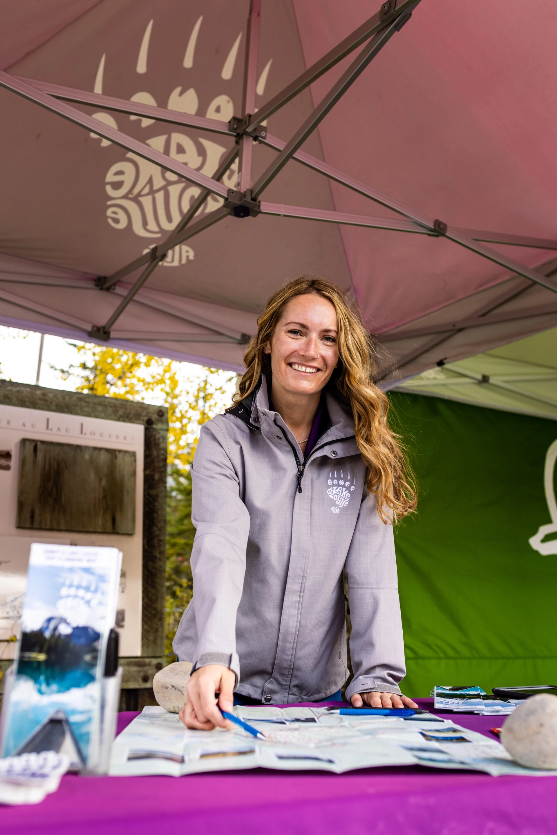 Person at outdoor info booth with brochures under a canopy tent near greenery.