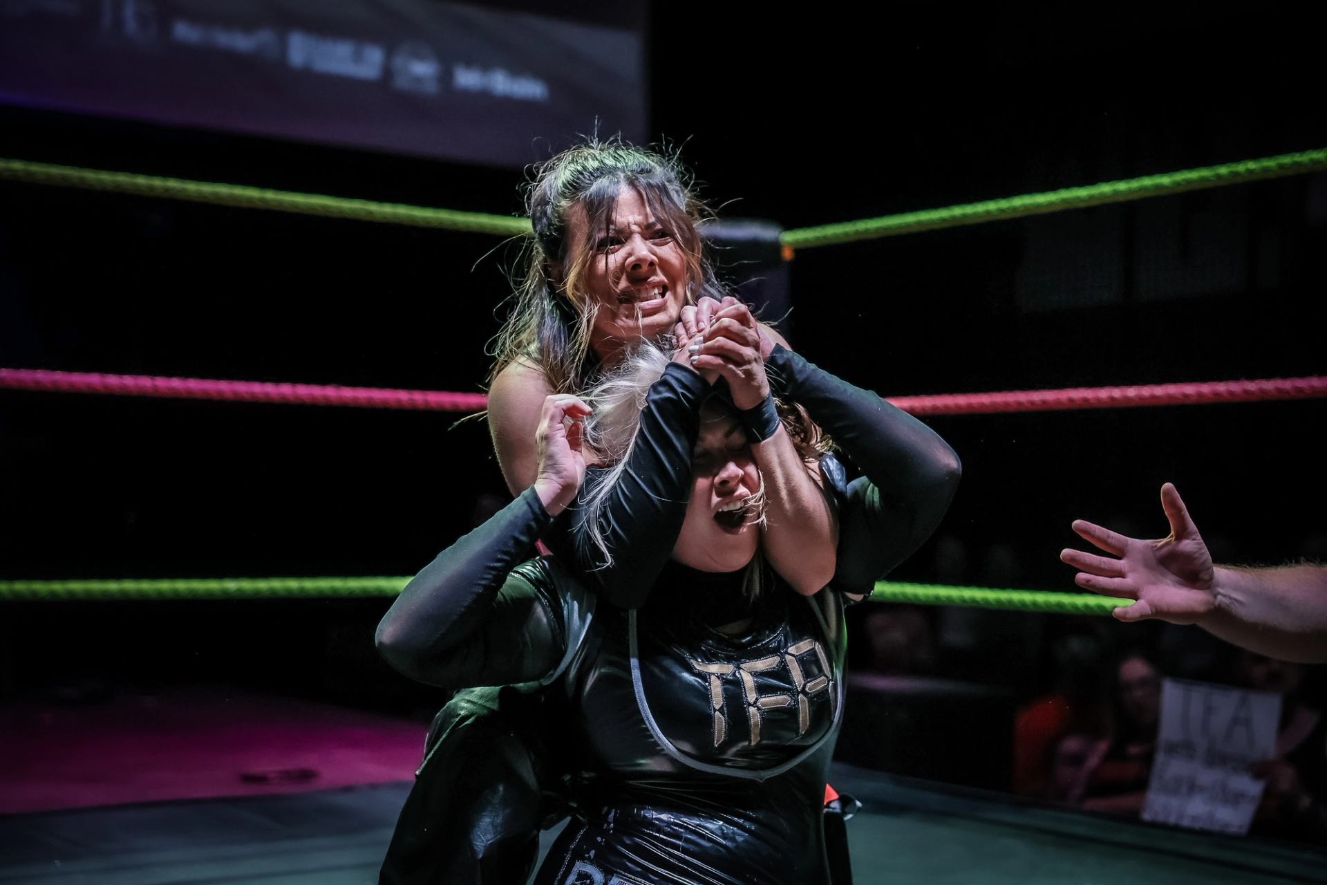 Professional wrestlers grapple in a hold inside a ring with colourful ropes.