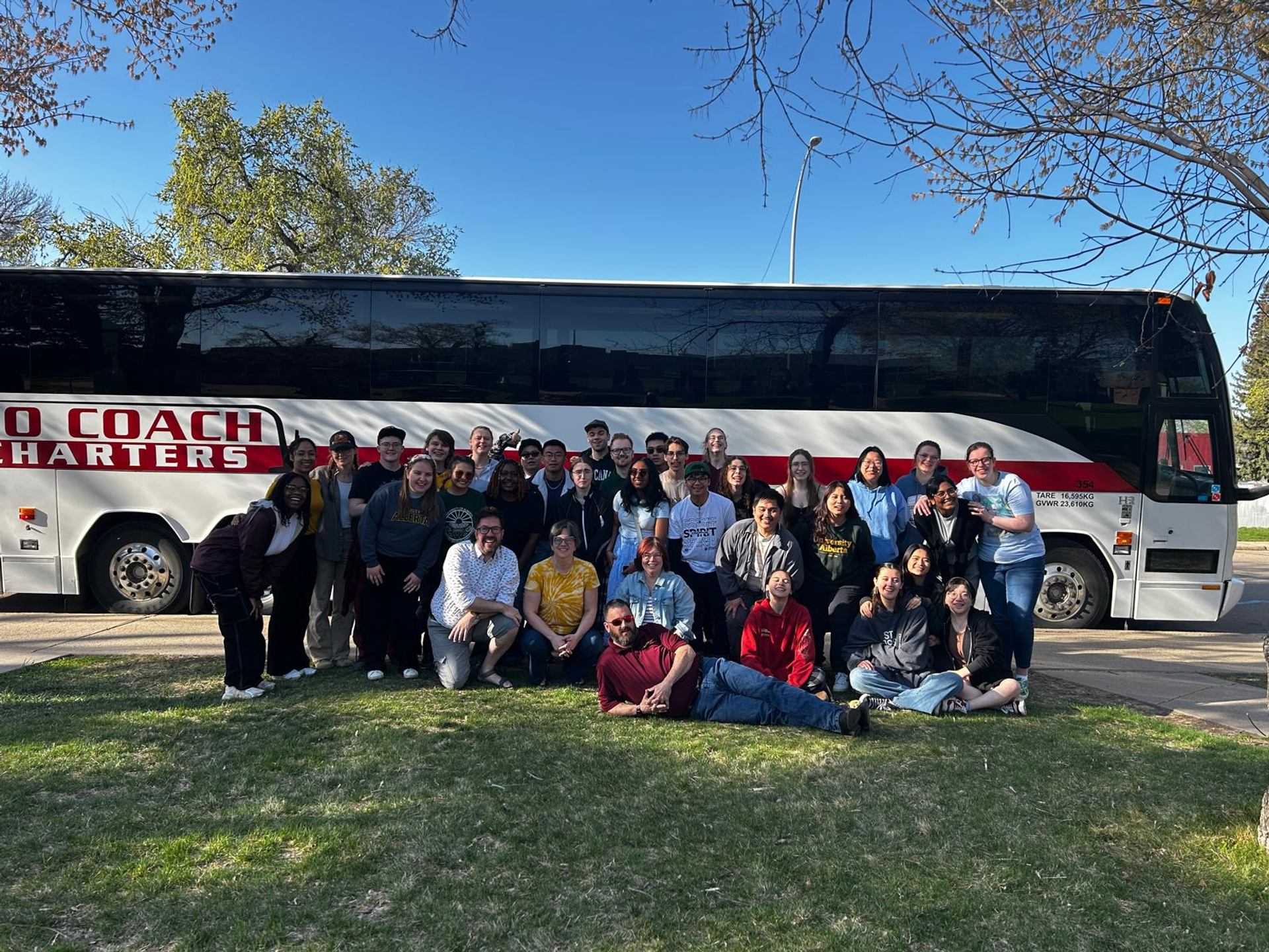 A group of people stands in front of a parked motorcoach on a grassy area beneath a clear blue sky.