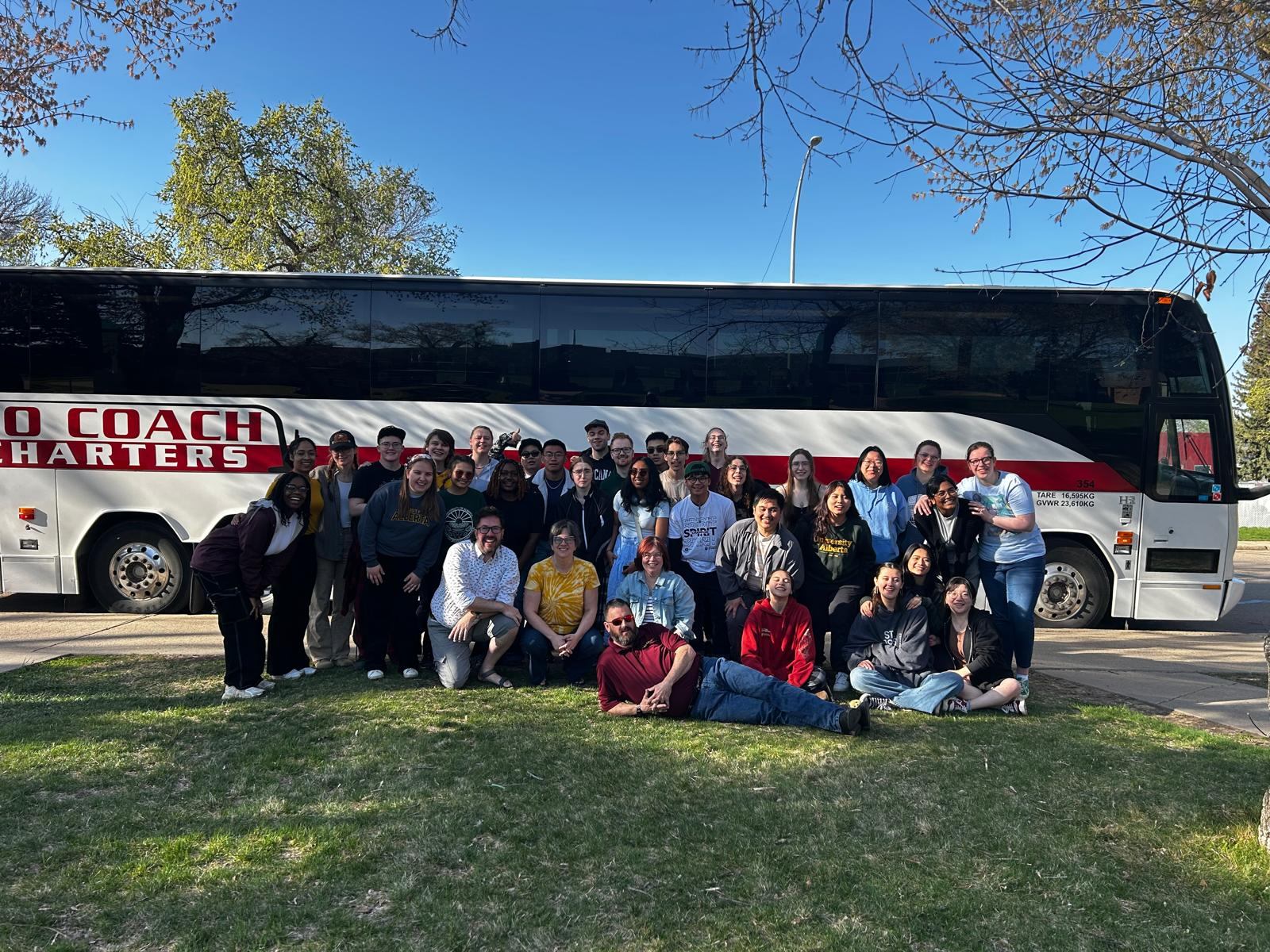 A group of people stands in front of a parked motorcoach on a grassy area beneath a clear blue sky.