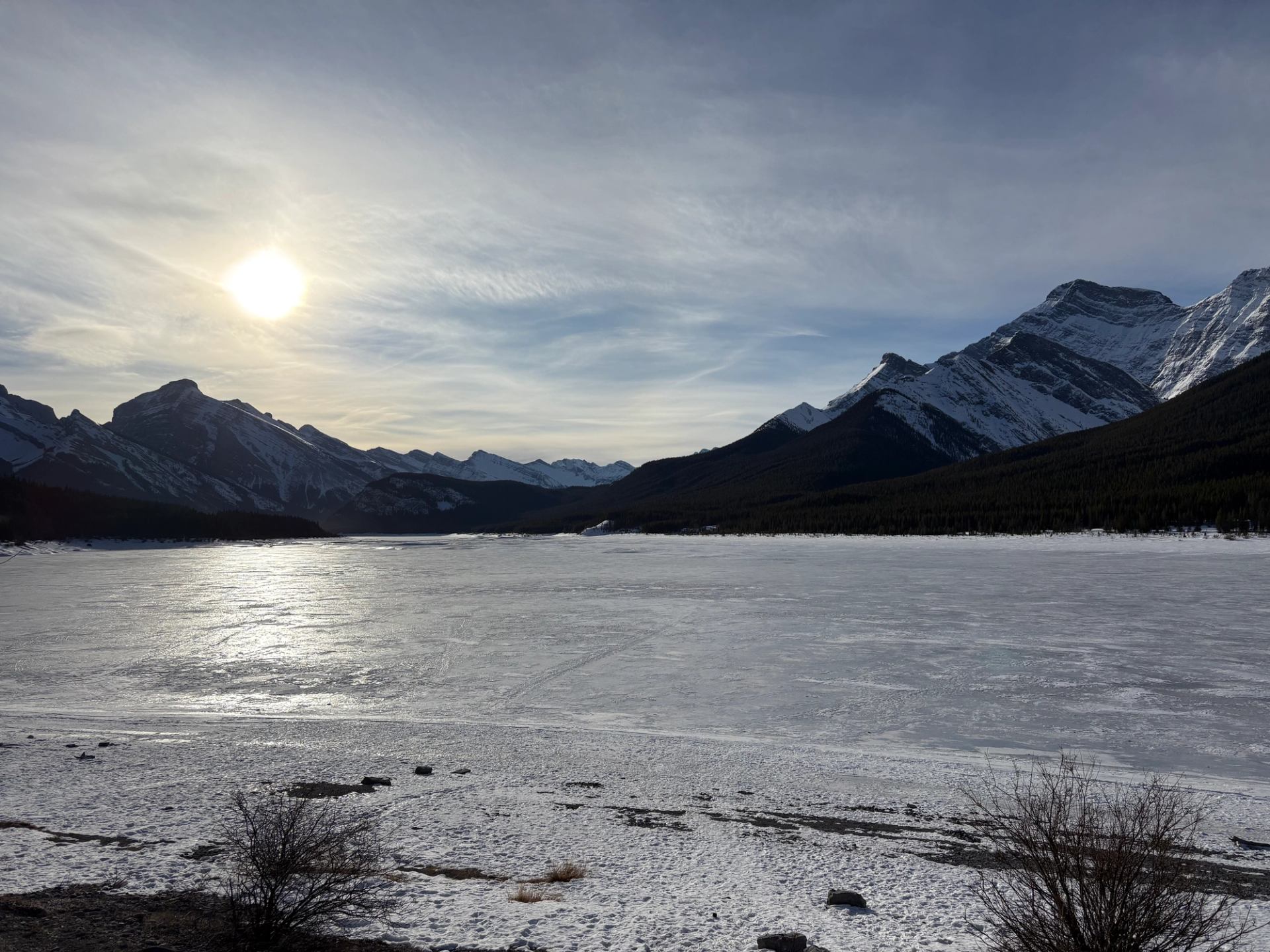 Frozen lake with snow-covered shoreline and distant mountain range under bright sun.