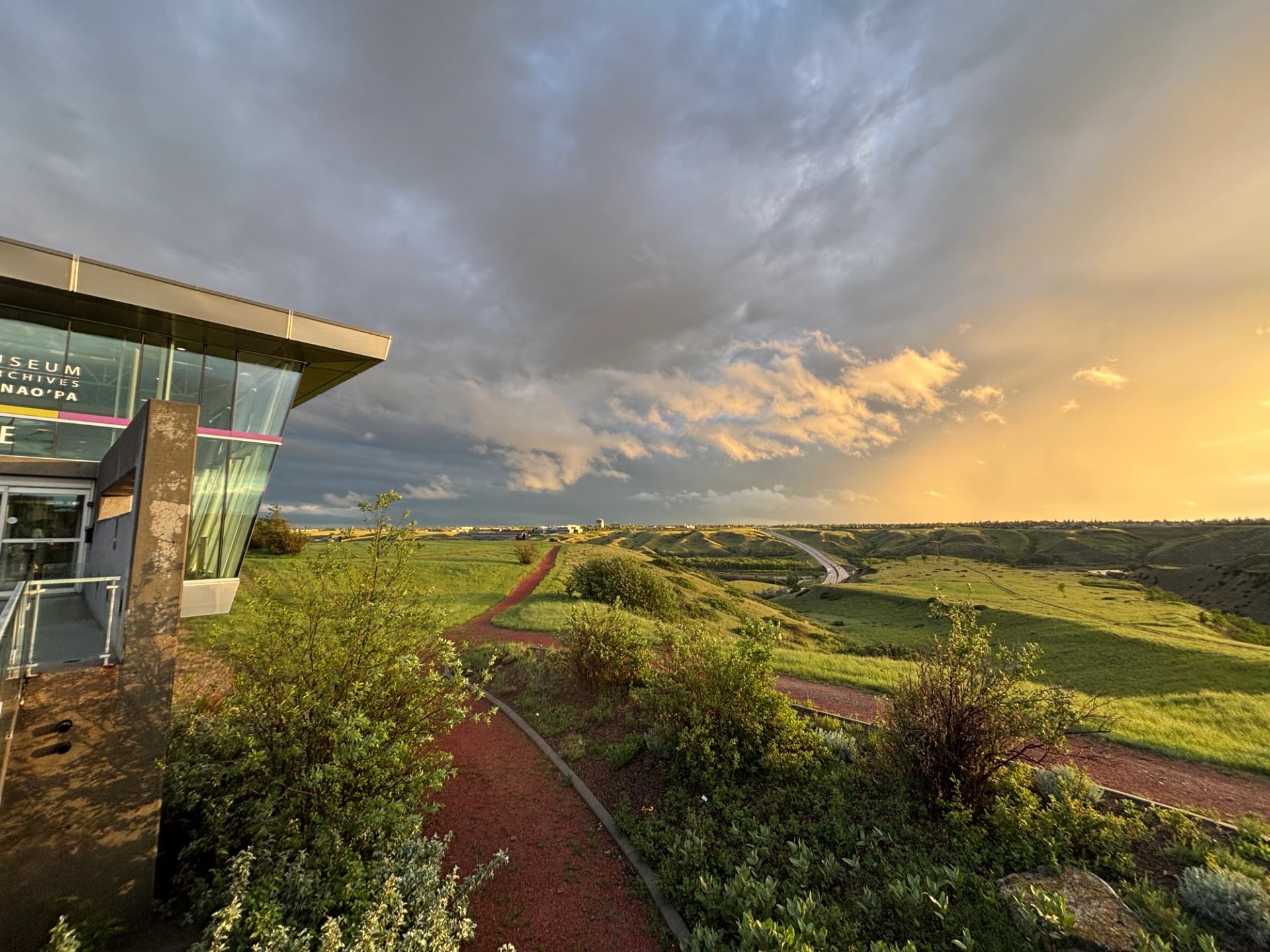 Glass museum building beside red gravel path overlooking green valley under dramatic sky.