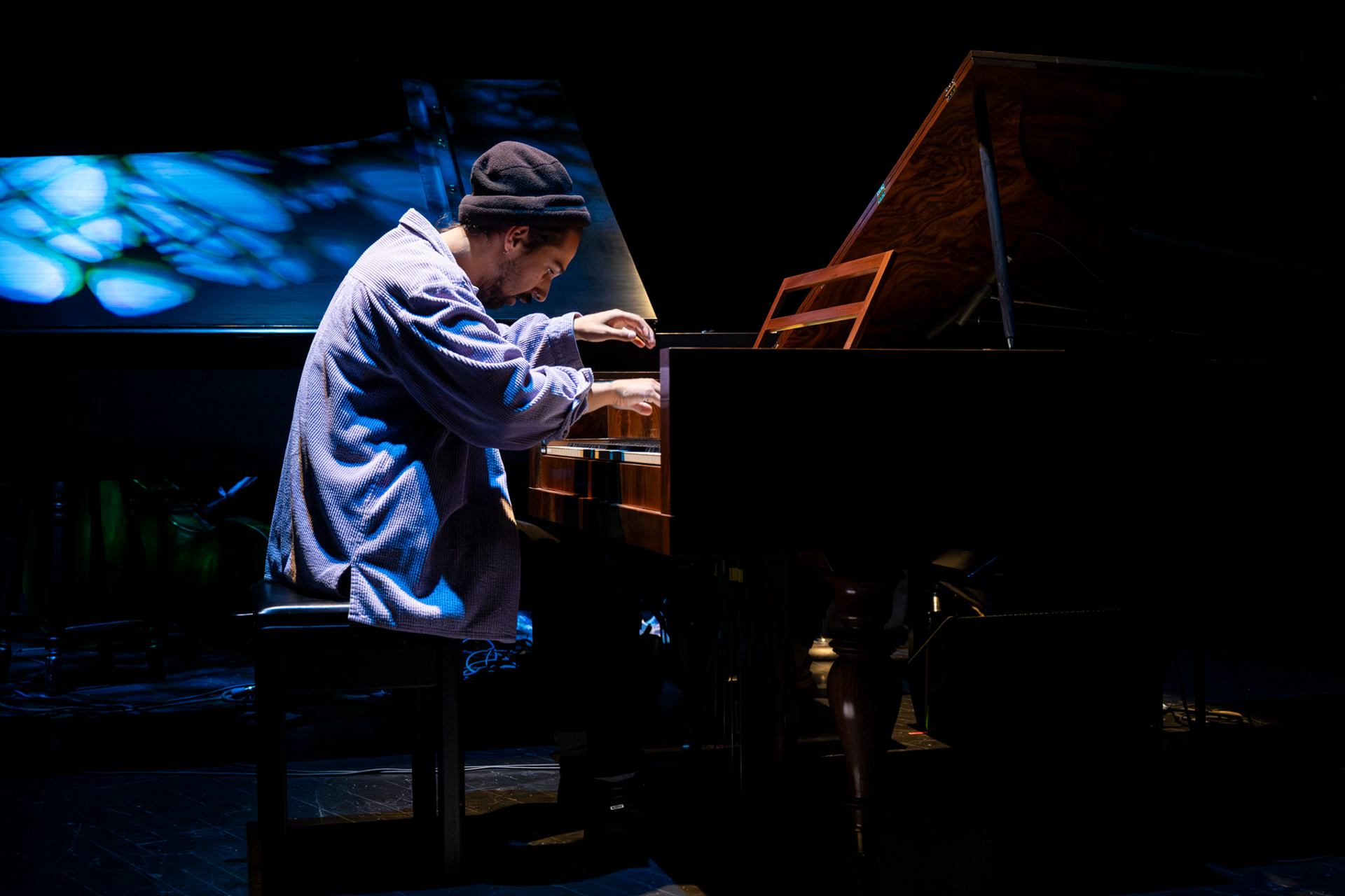 Pianist in loose shirt and toque plays grand piano on dark stage under blue light.